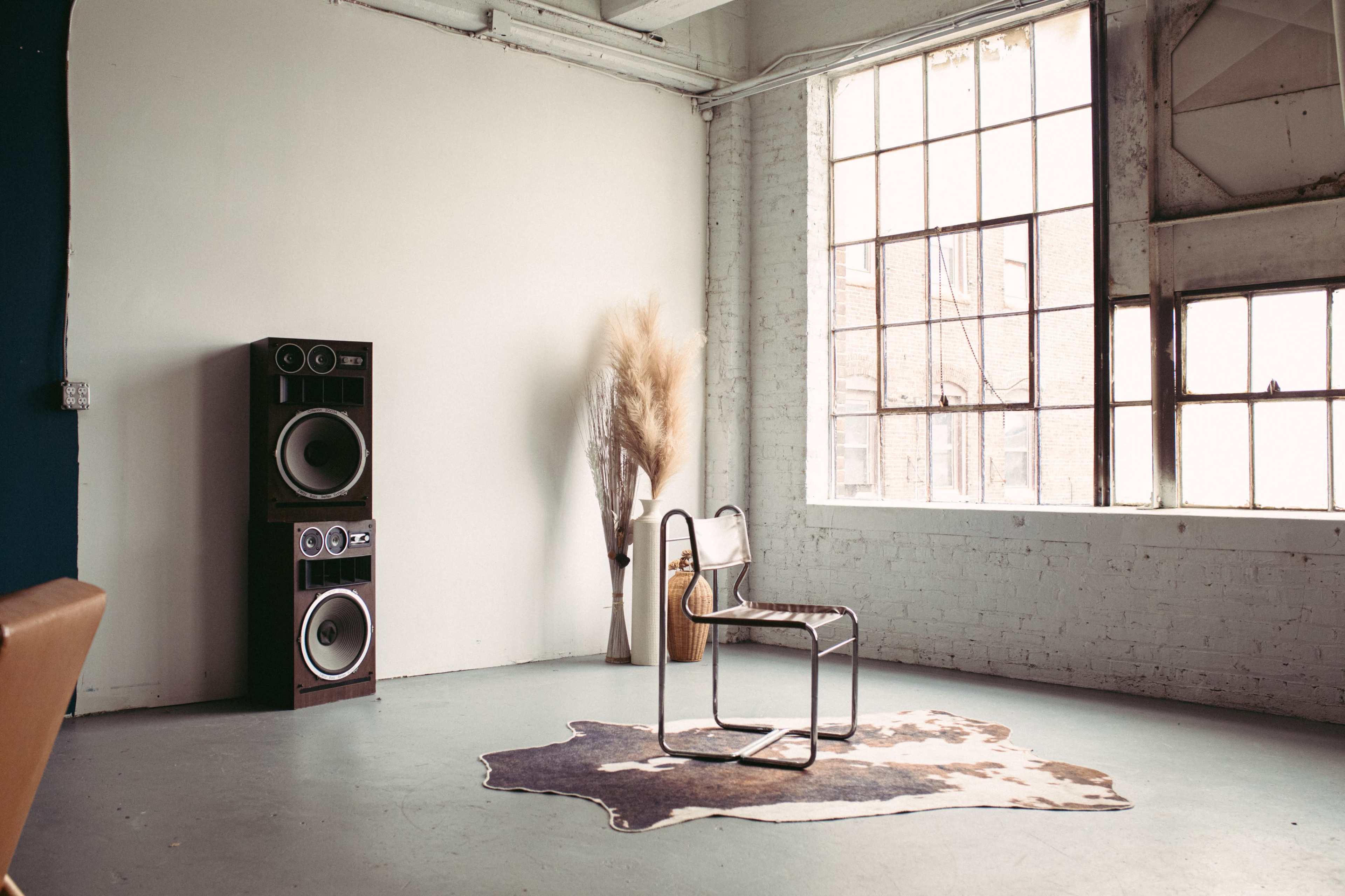 The image shows a minimalist room with a single chair on a cowhide rug, a tall speaker set against the wall, and a large window letting in natural light.