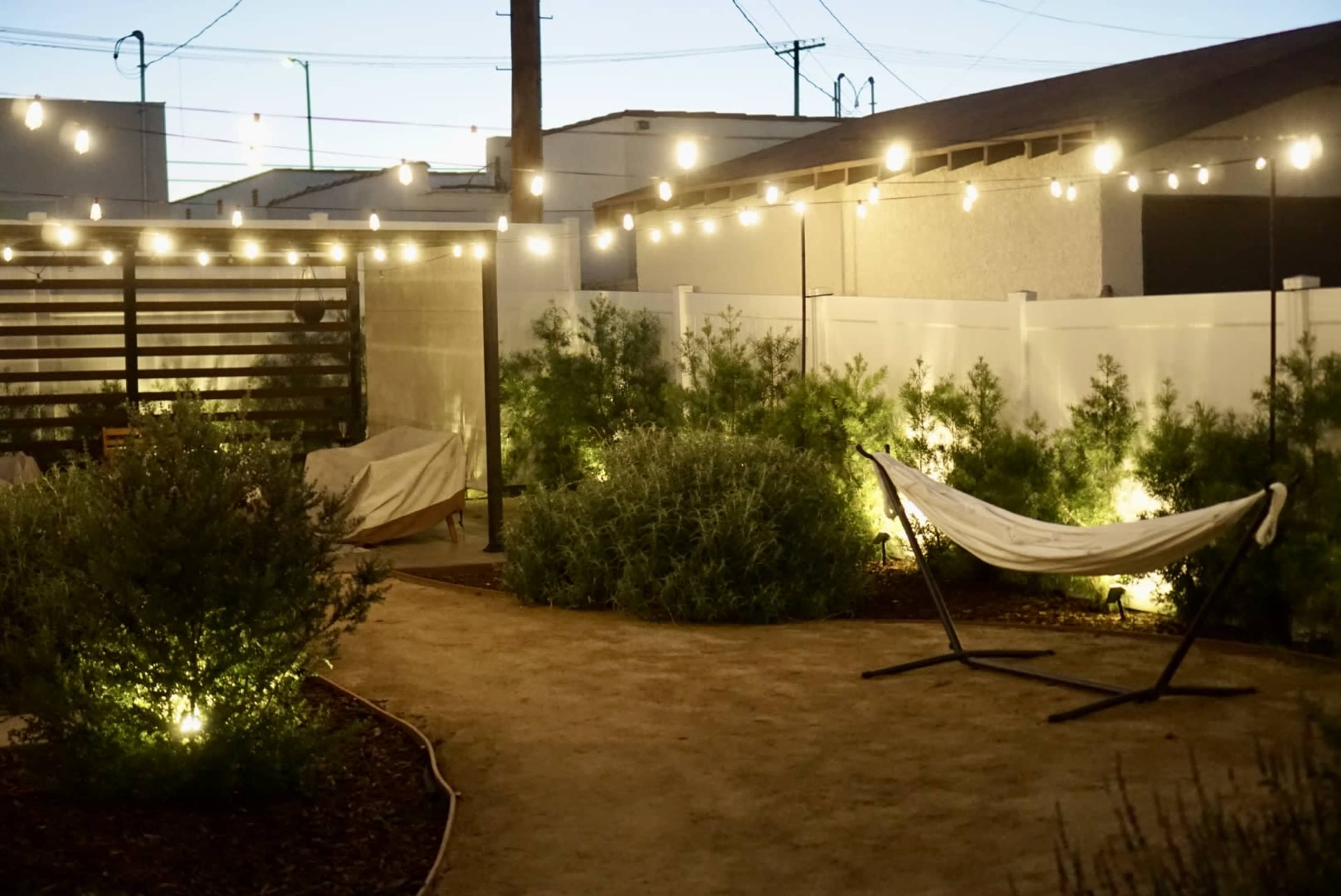 A well-lit outdoor space features a hammock, surrounded by low bushes and string lights overhead.