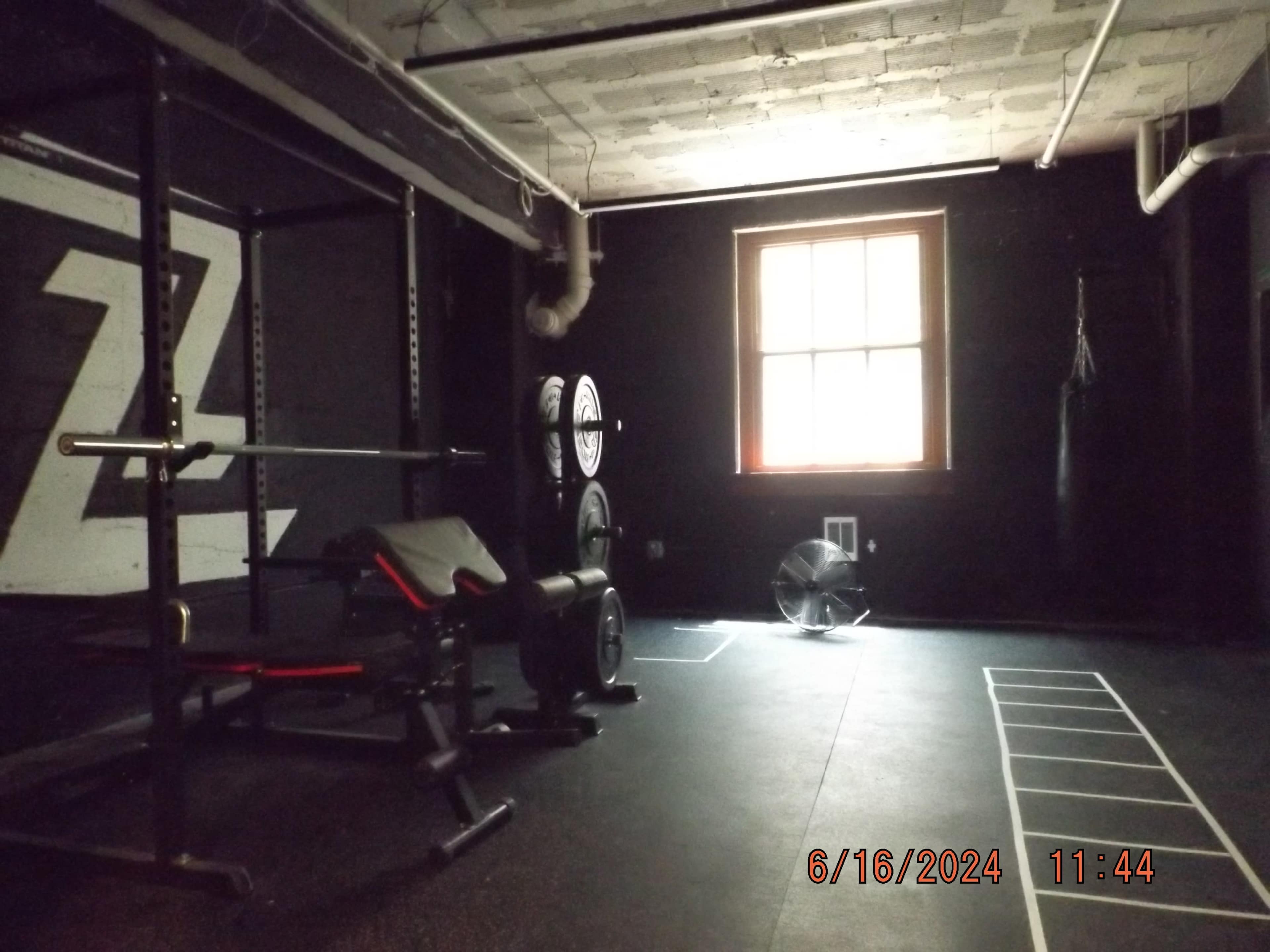 The image shows a dimly lit gym with a squat rack, weights, a bench, and a fan, alongside a wall marked with a large letter "Z."