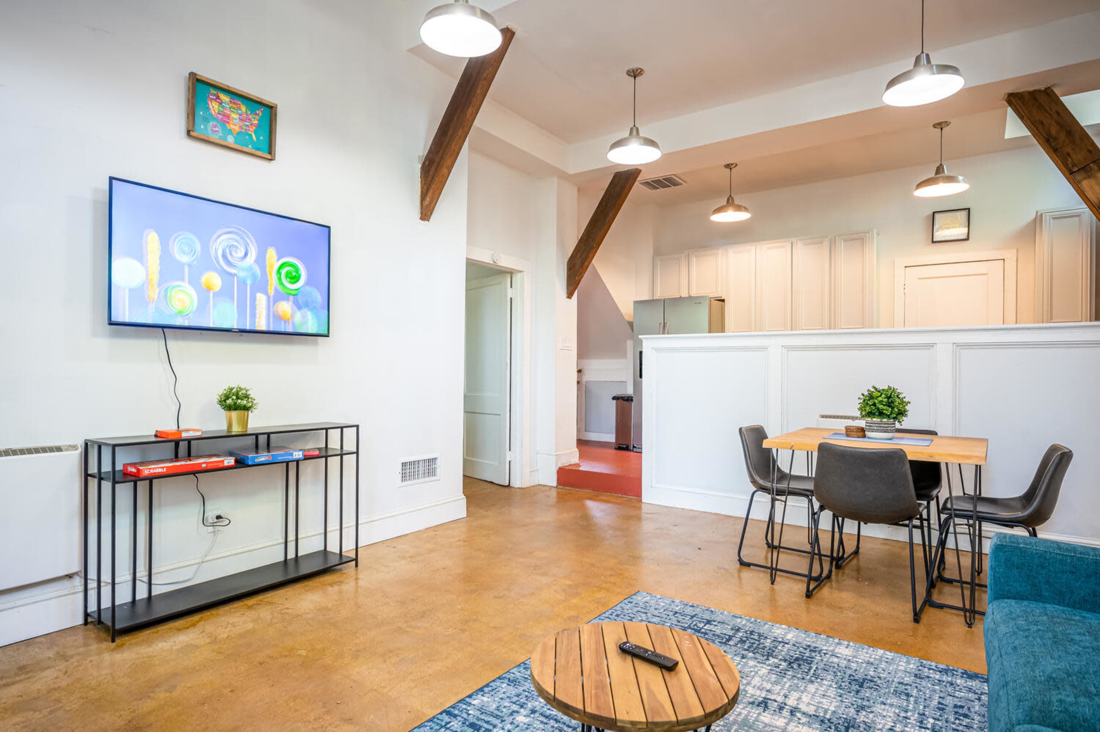 The living space features a television mounted on a black stand, a blue rug, a dining area with a wooden table and chairs, and visible kitchen appliances in the background.