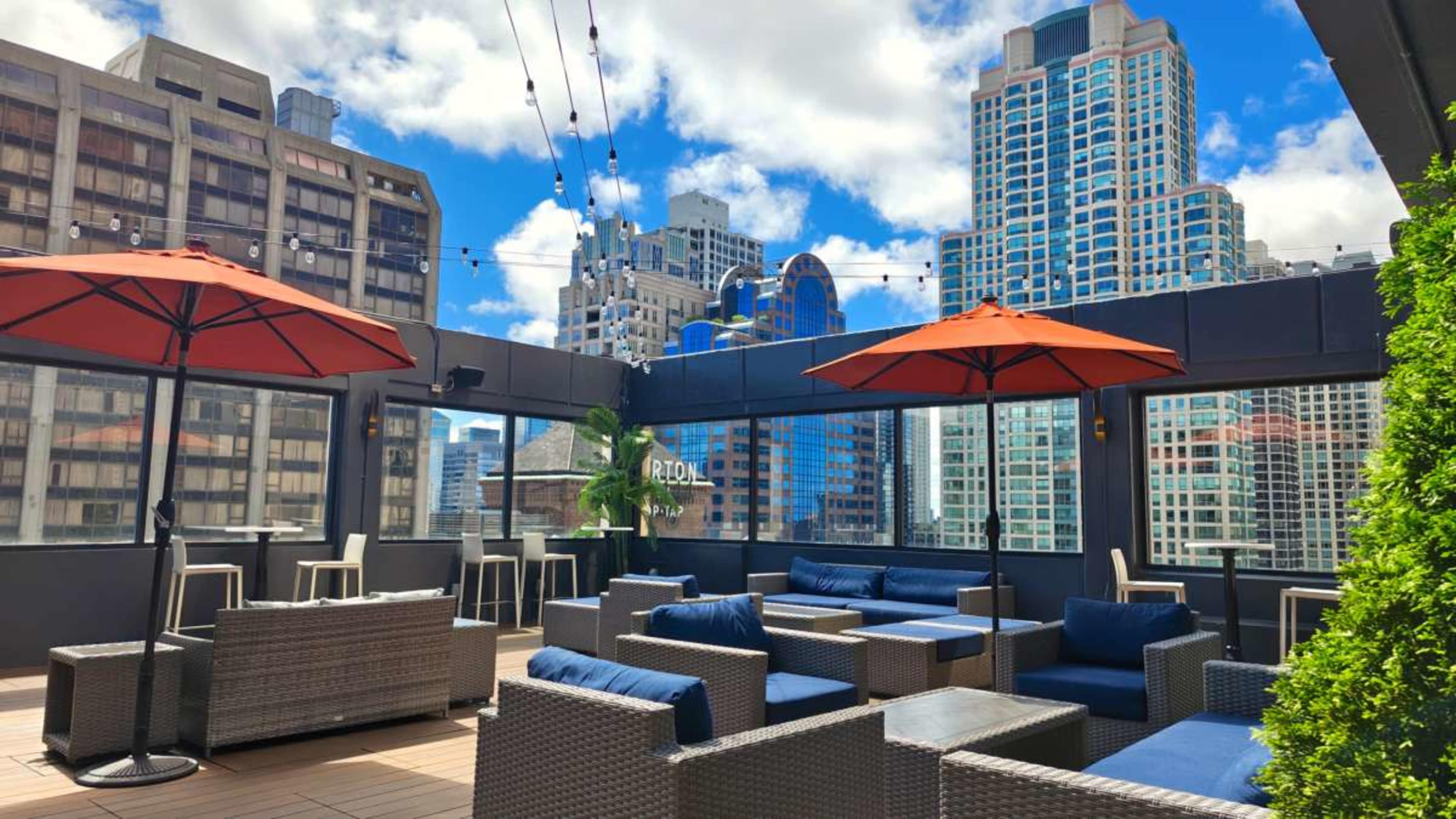 The image shows a rooftop lounge area with wicker furniture and orange umbrellas, situated among tall urban buildings under a partly cloudy sky.