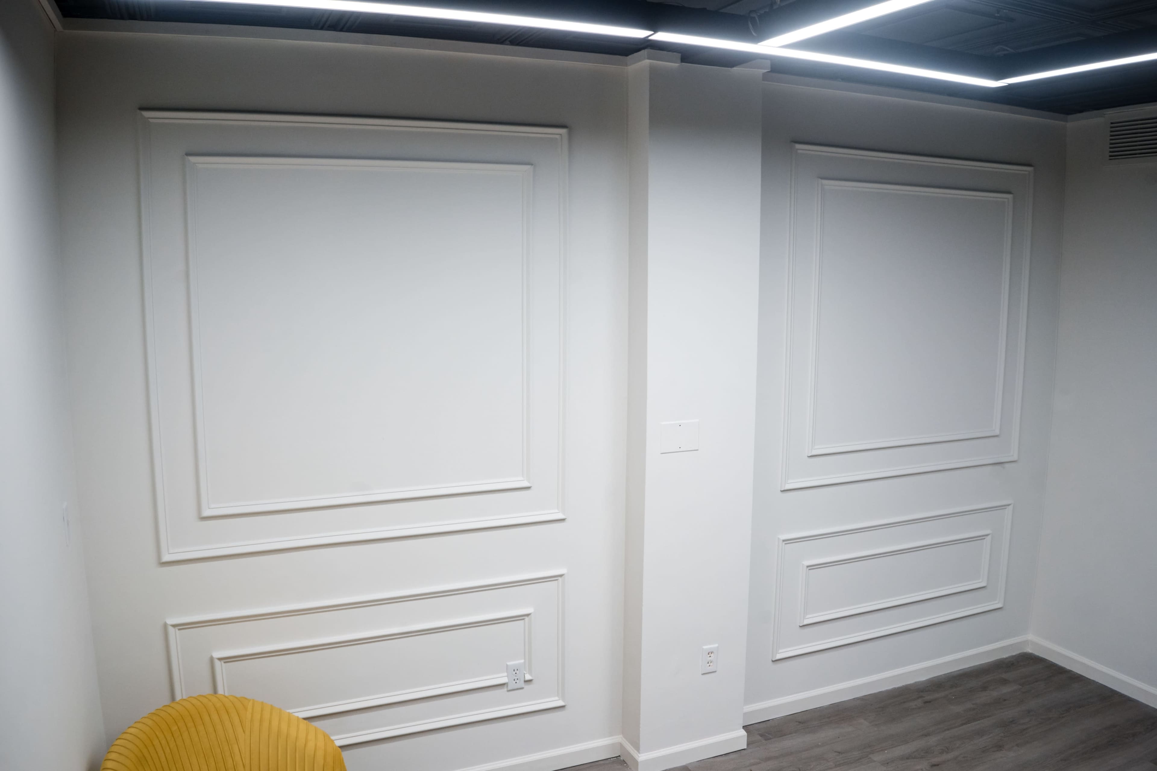The image shows a minimalistic room with white walls featuring decorative molding and a yellow chair in the foreground.