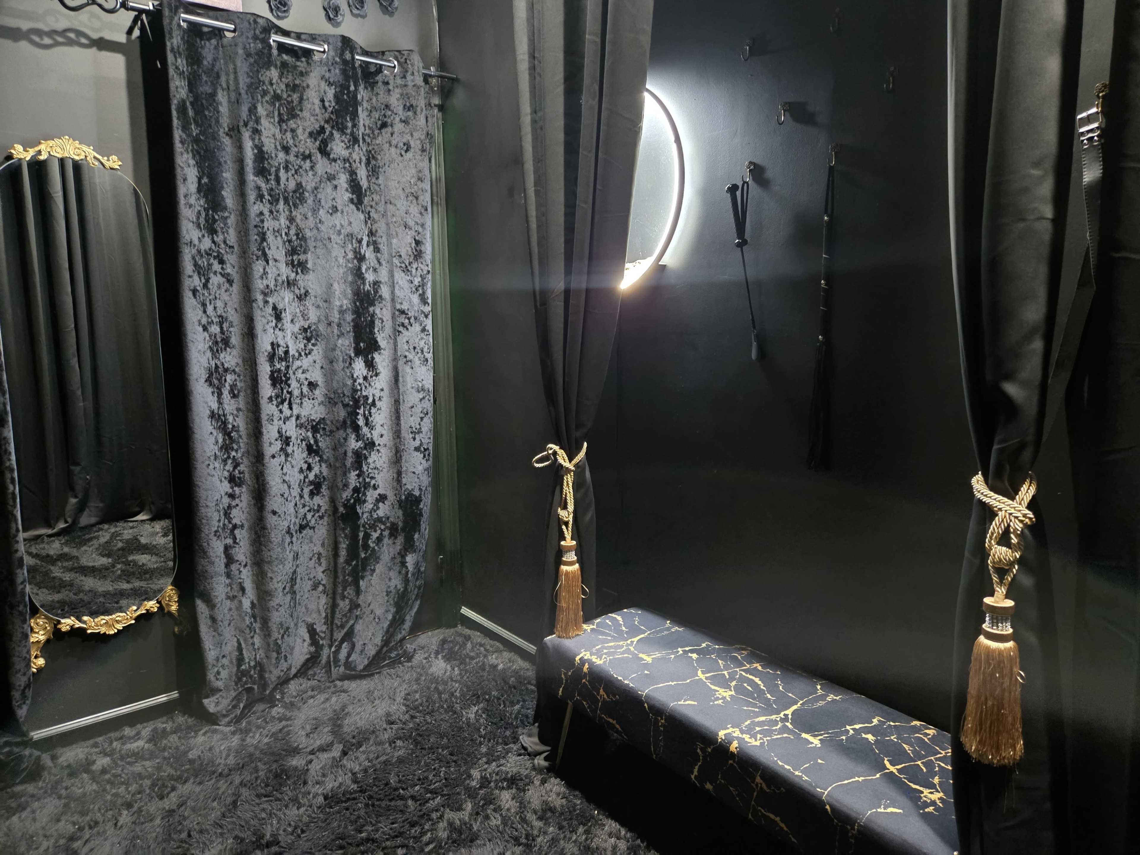The image shows a dimly lit room with black velvet curtains, a circular mirror, and a decorative bench with gold accents.