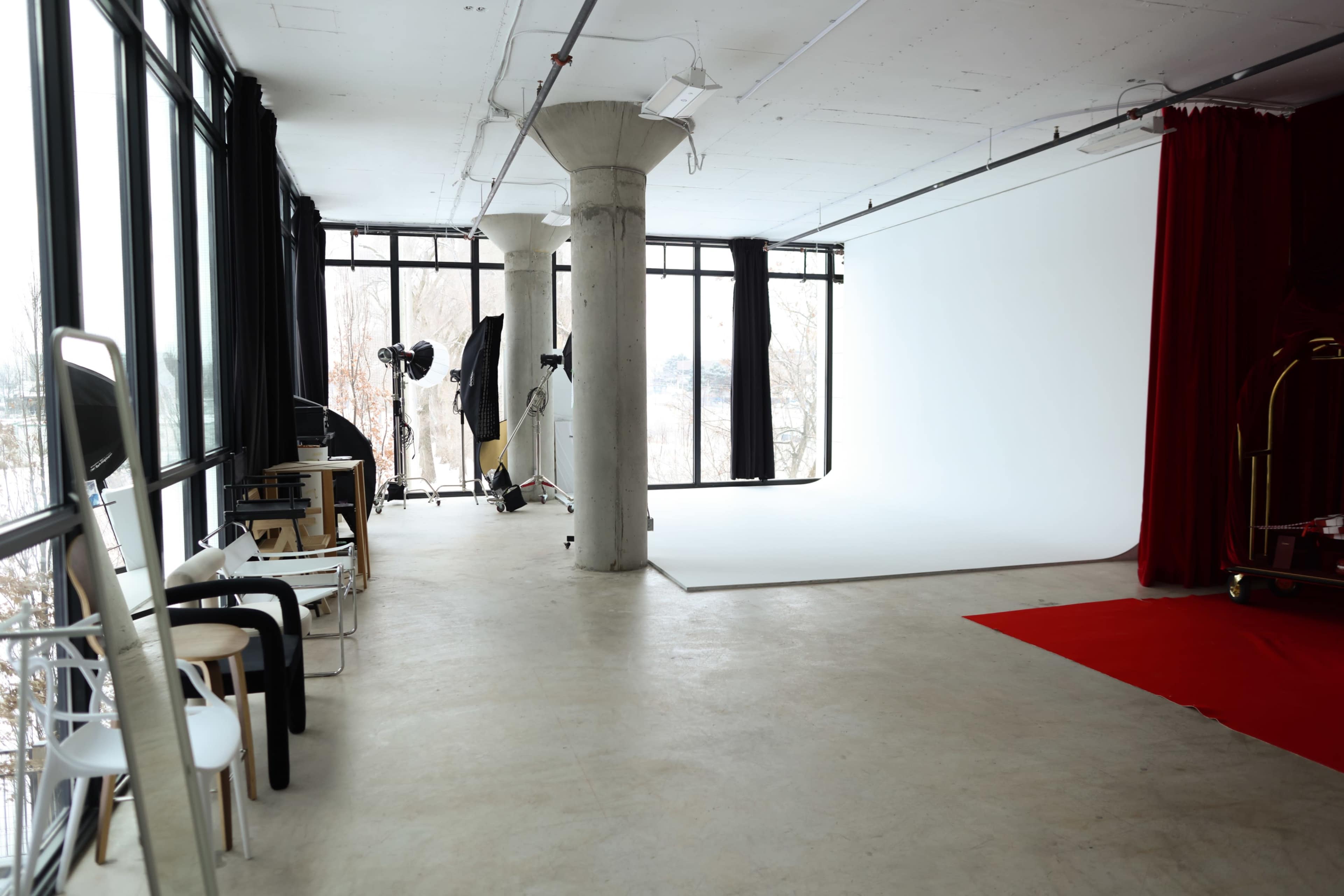 The image shows a spacious photography studio featuring large windows, a white backdrop, several chairs, and a red carpet area.