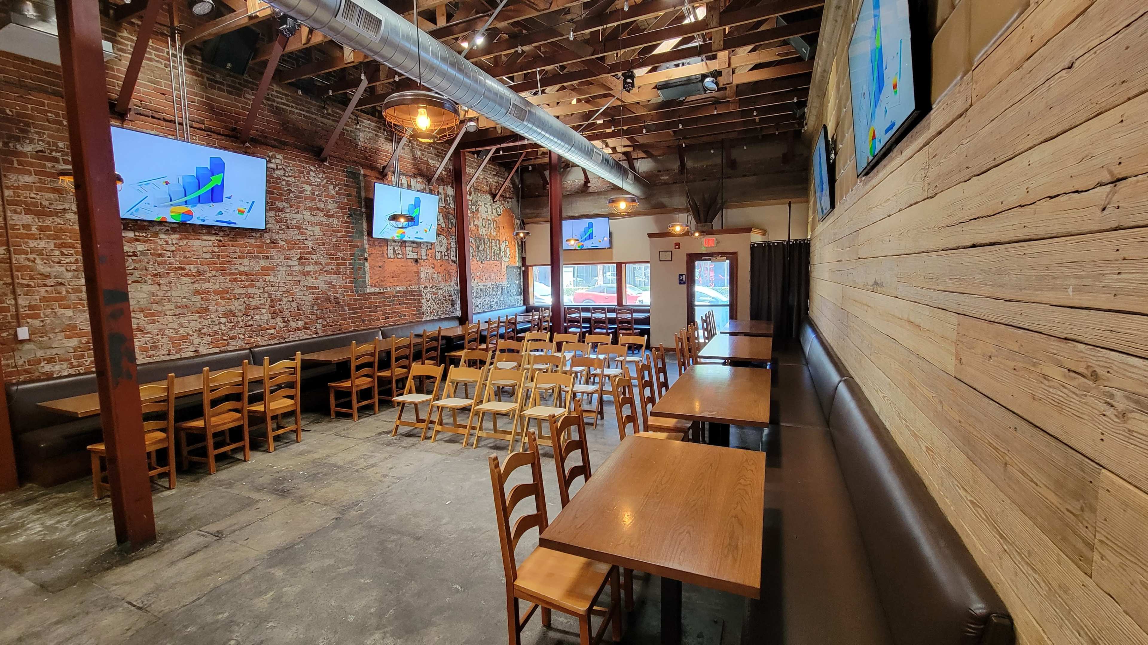 A spacious restaurant interior with wooden tables and chairs set up in rows, exposed brick walls, and several screens mounted on the walls.