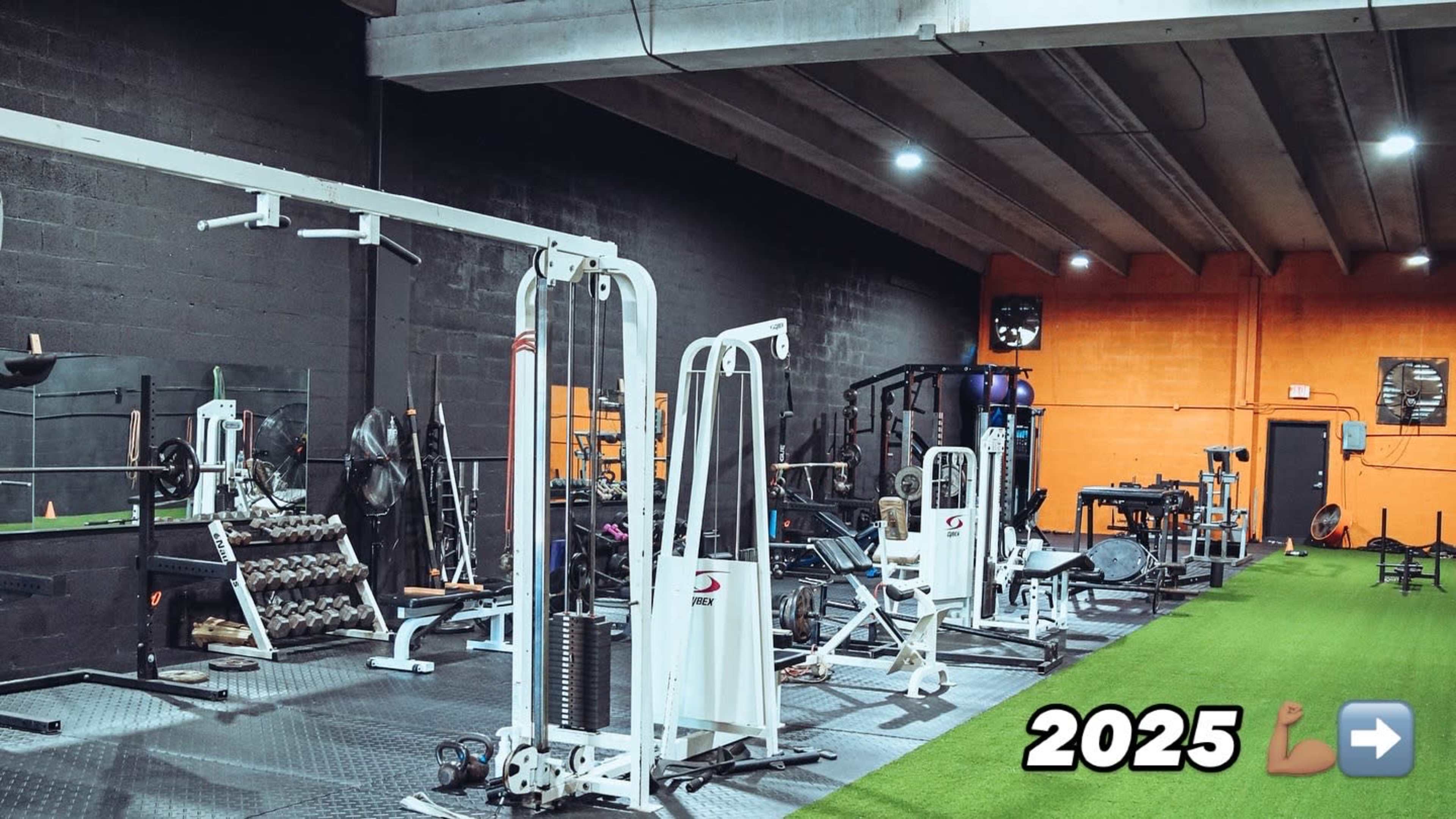 The image shows a gym interior equipped with various exercise machines and weights, featuring a turf area and a bright orange wall.