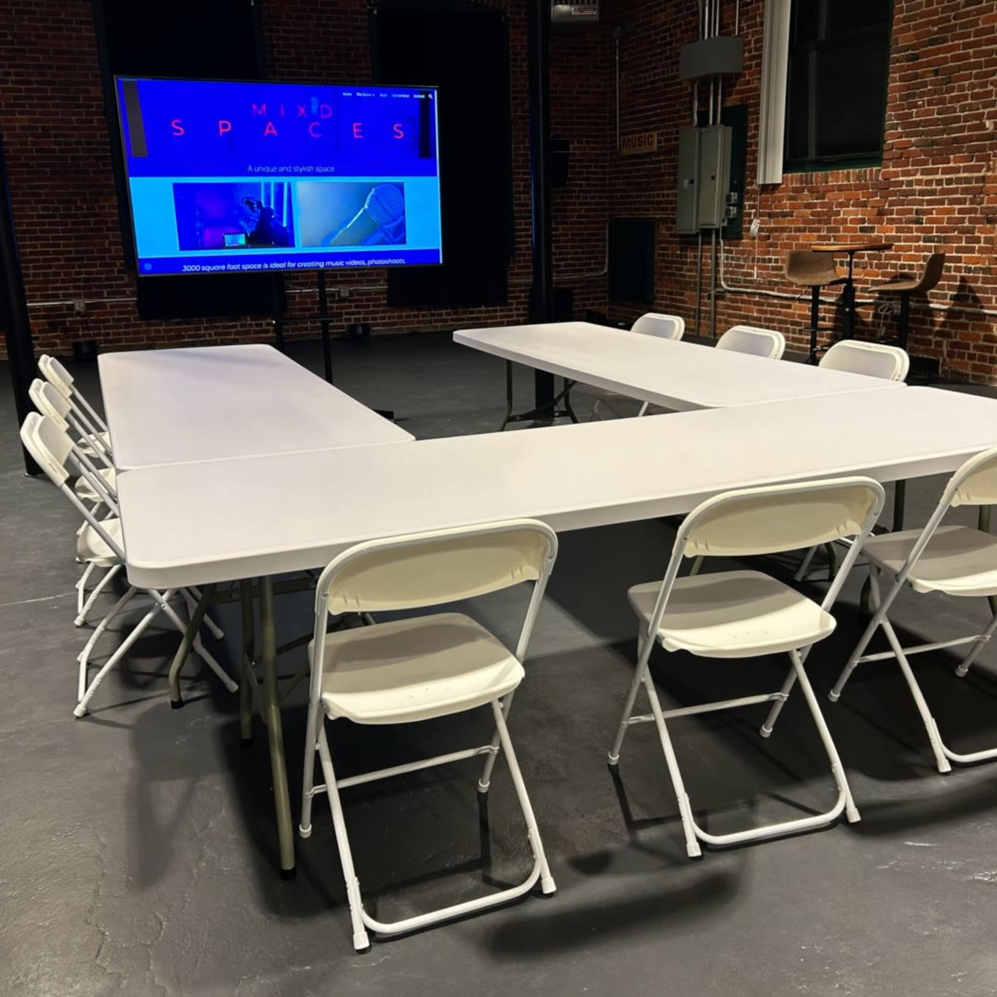 A spacious meeting room features a U-shaped arrangement of white tables and chairs, with a large screen displaying a digital presentation in the background.