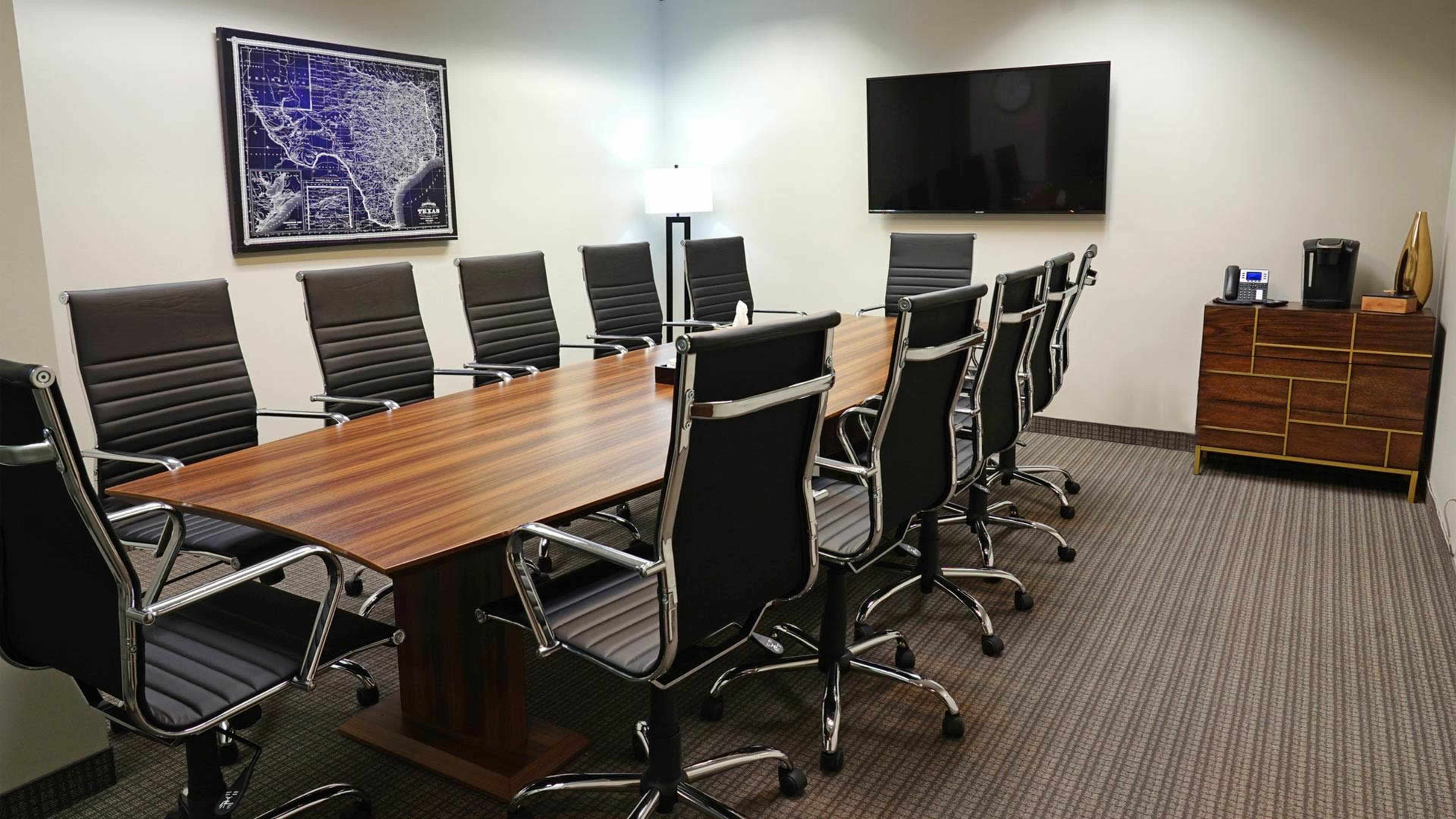 The image shows a modern conference room with a long wooden table surrounded by black ergonomic chairs, and equipped with a wall-mounted television and a coffee machine on a side cabinet.