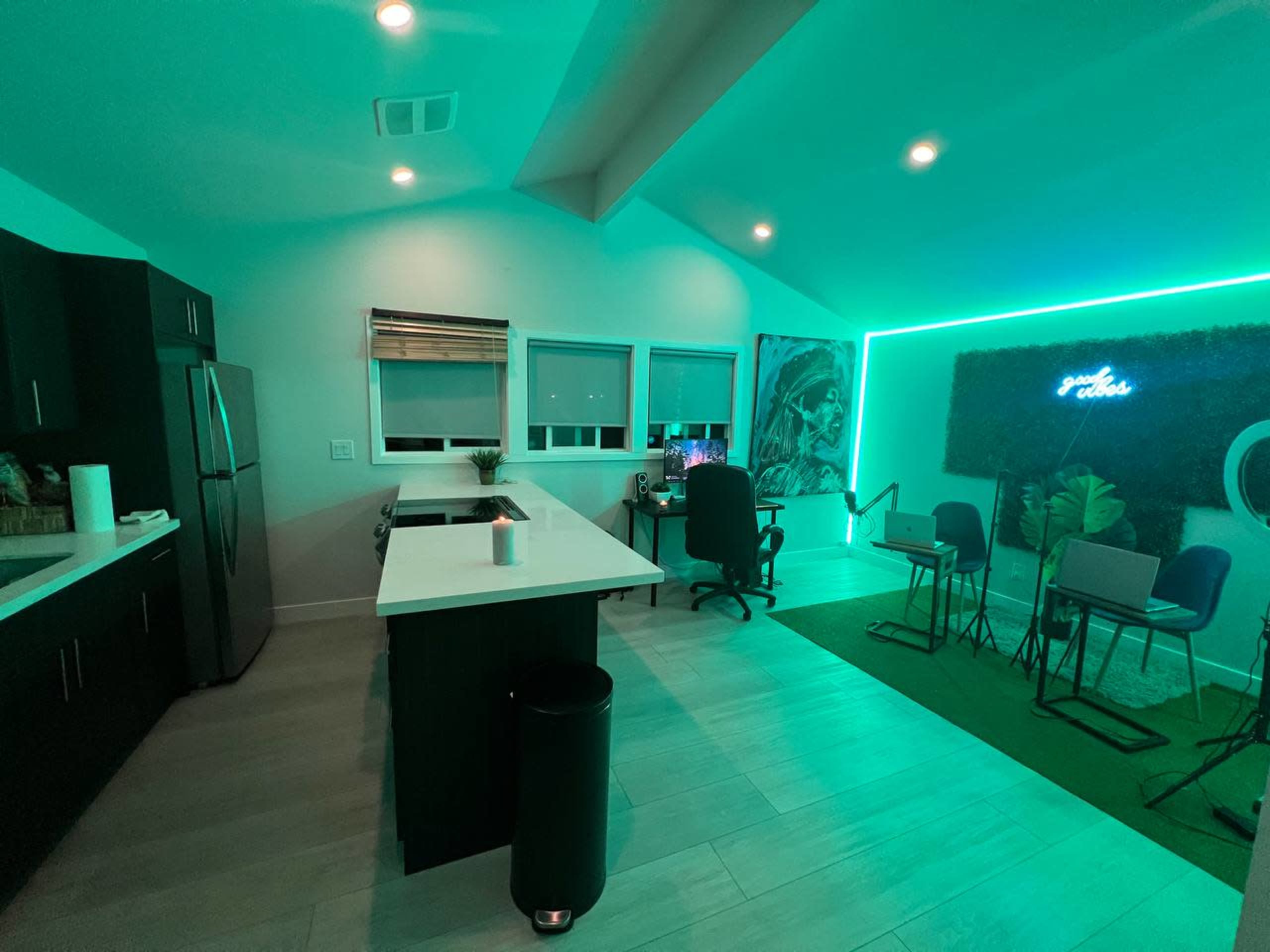 A modern kitchen and workspace area illuminated with green LED lighting, featuring a kitchen island, a desk with a computer chair, and a small seating arrangement.