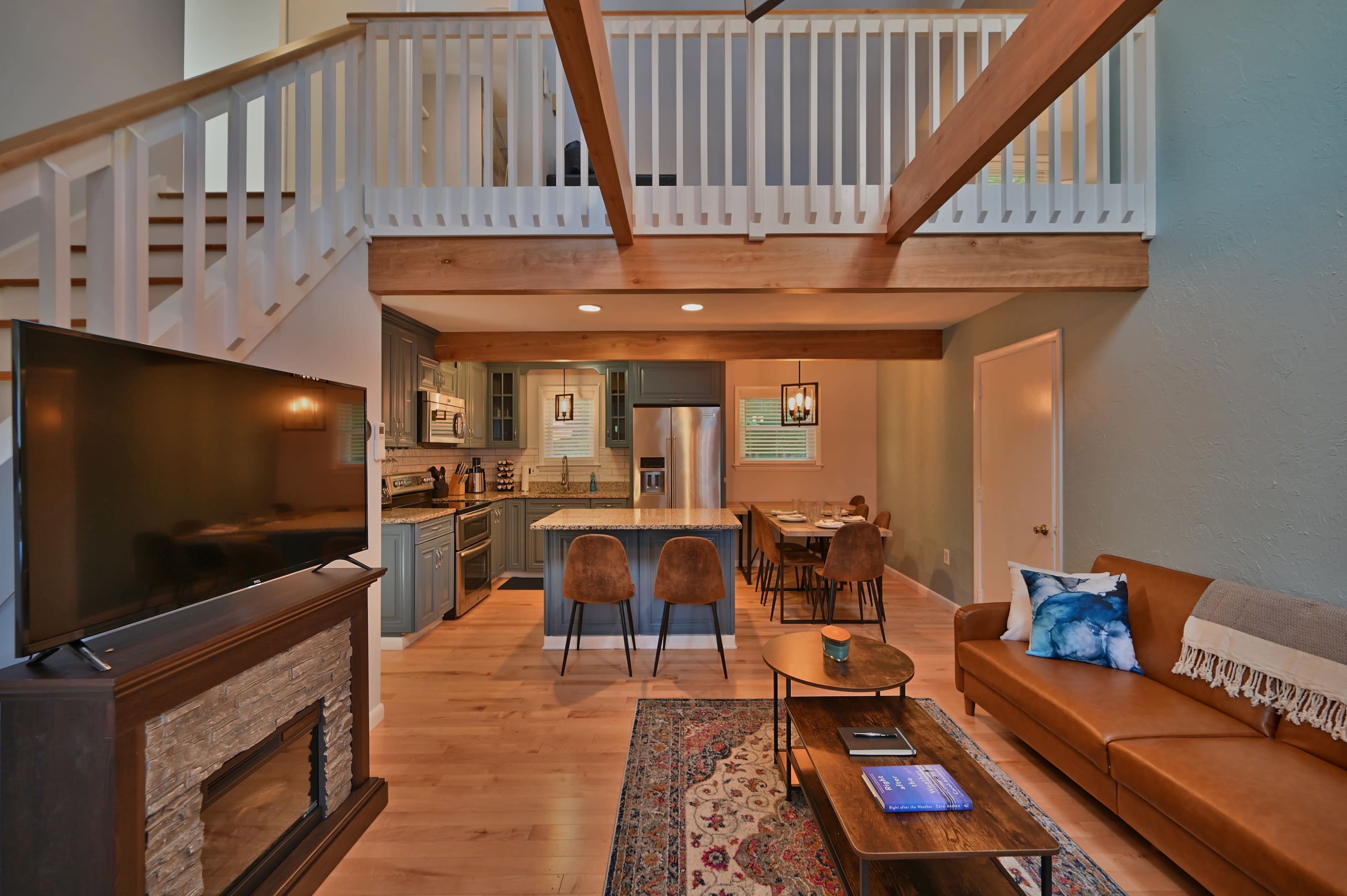 The image shows a modern living space with a staircase, an open kitchen area, a living room featuring a leather couch, and wooden beams across the ceiling.