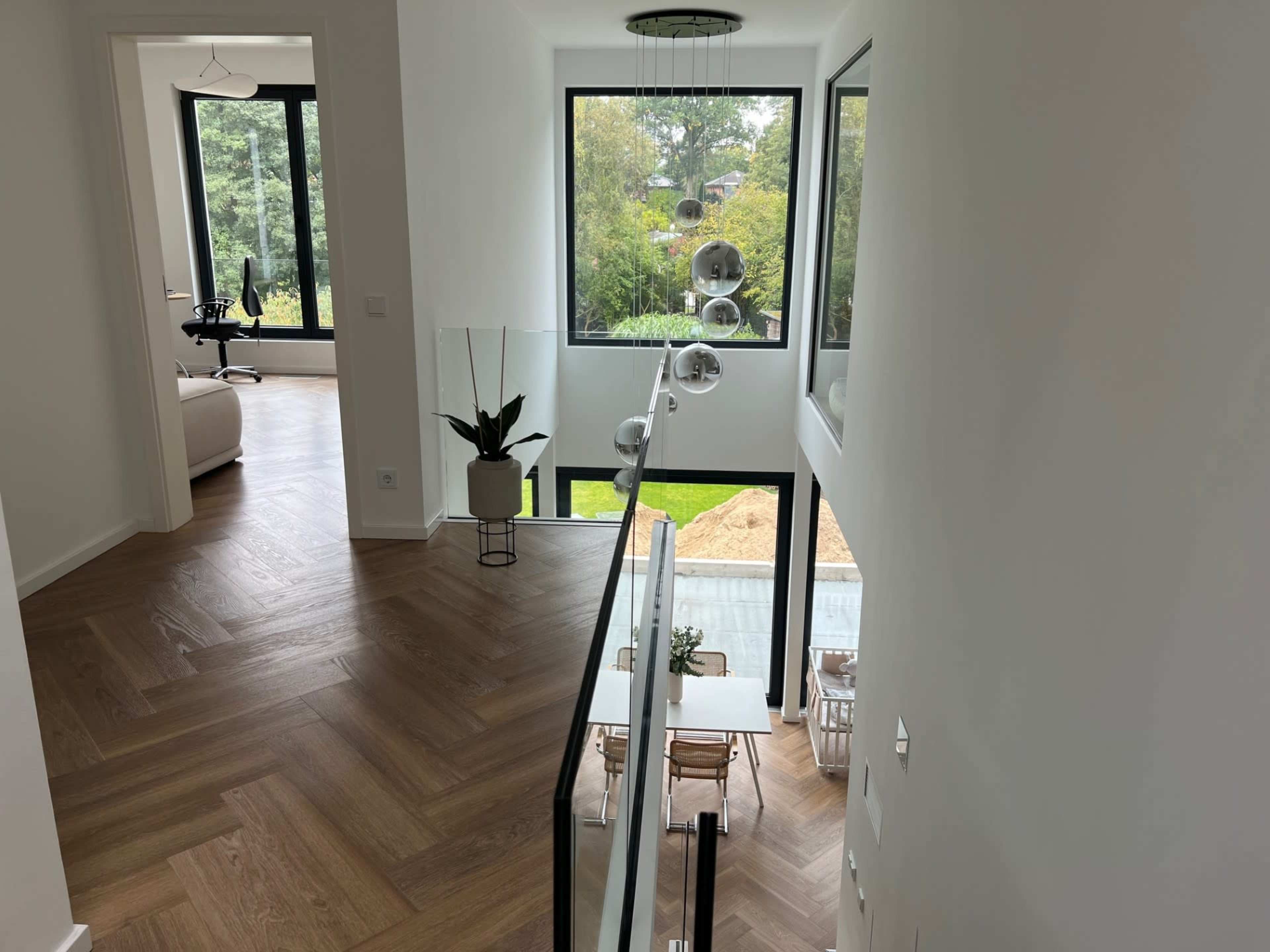 A modern hallway features large windows overlooking a garden and a dining area below, with wood flooring and minimal decor.