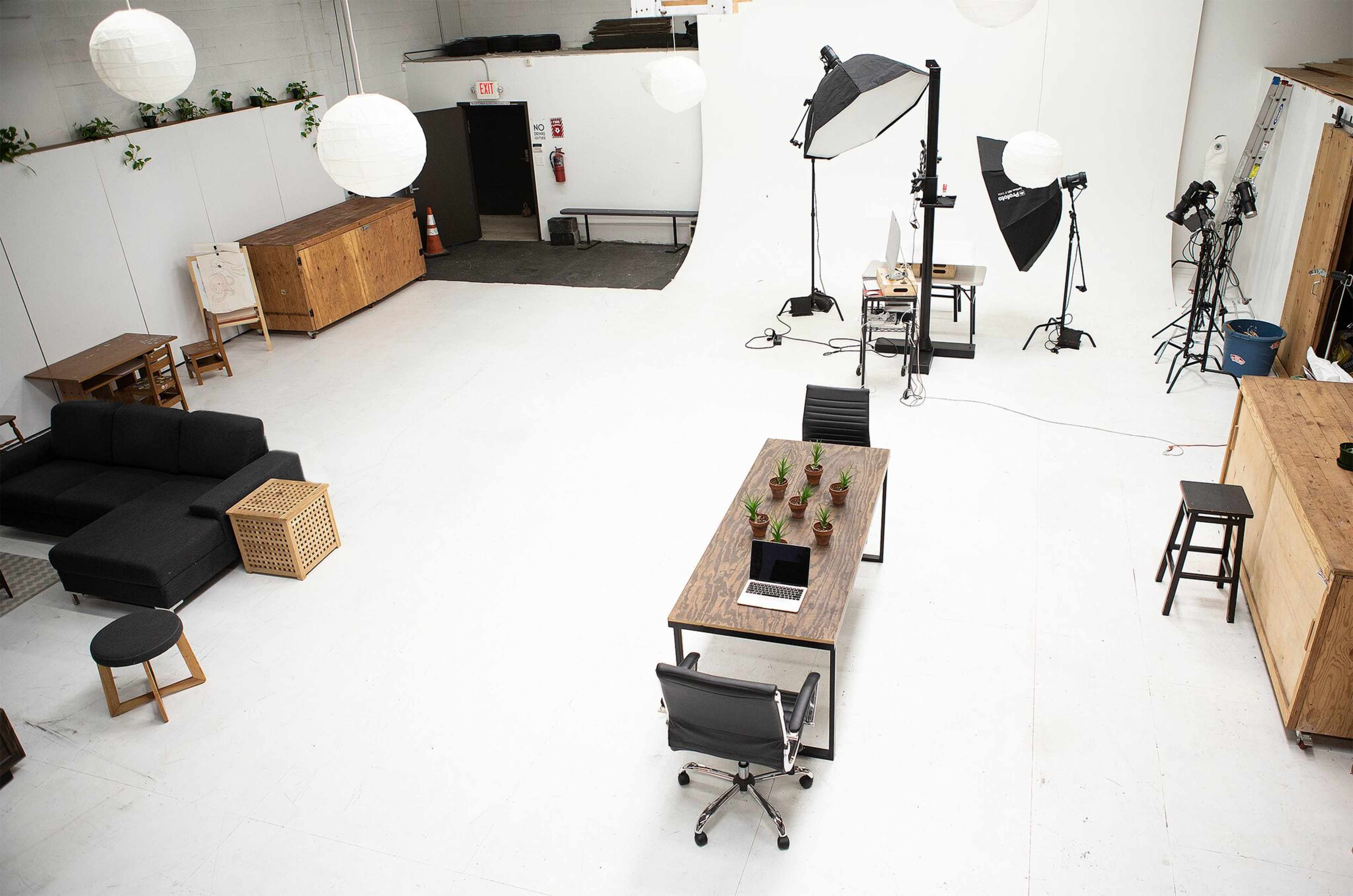 A spacious studio with a white backdrop, featuring a large wooden table, several potted plants, and various photography equipment.
