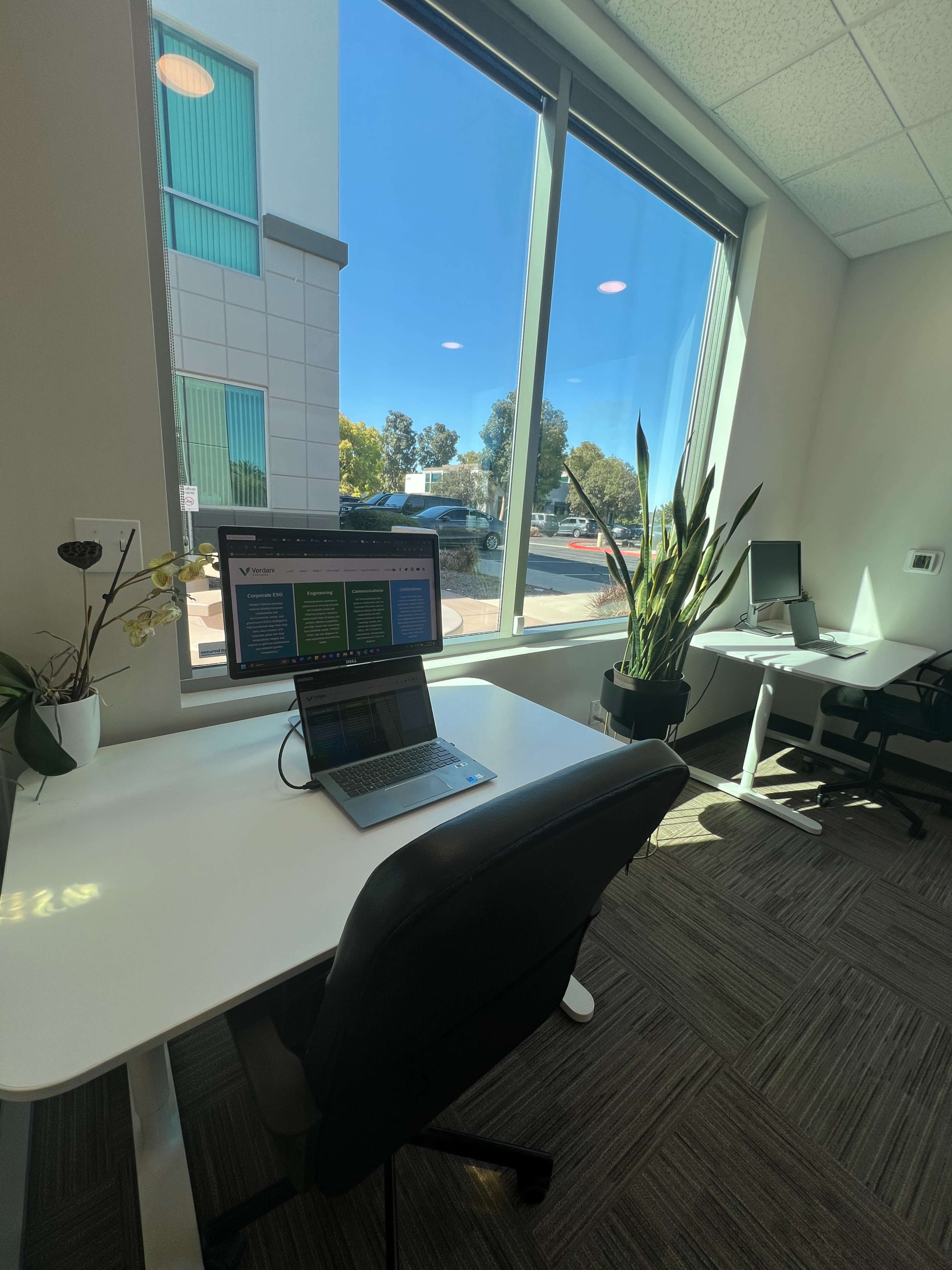 A bright office space features two desks, one with a laptop open, and large windows overlooking a parking lot and greenery.