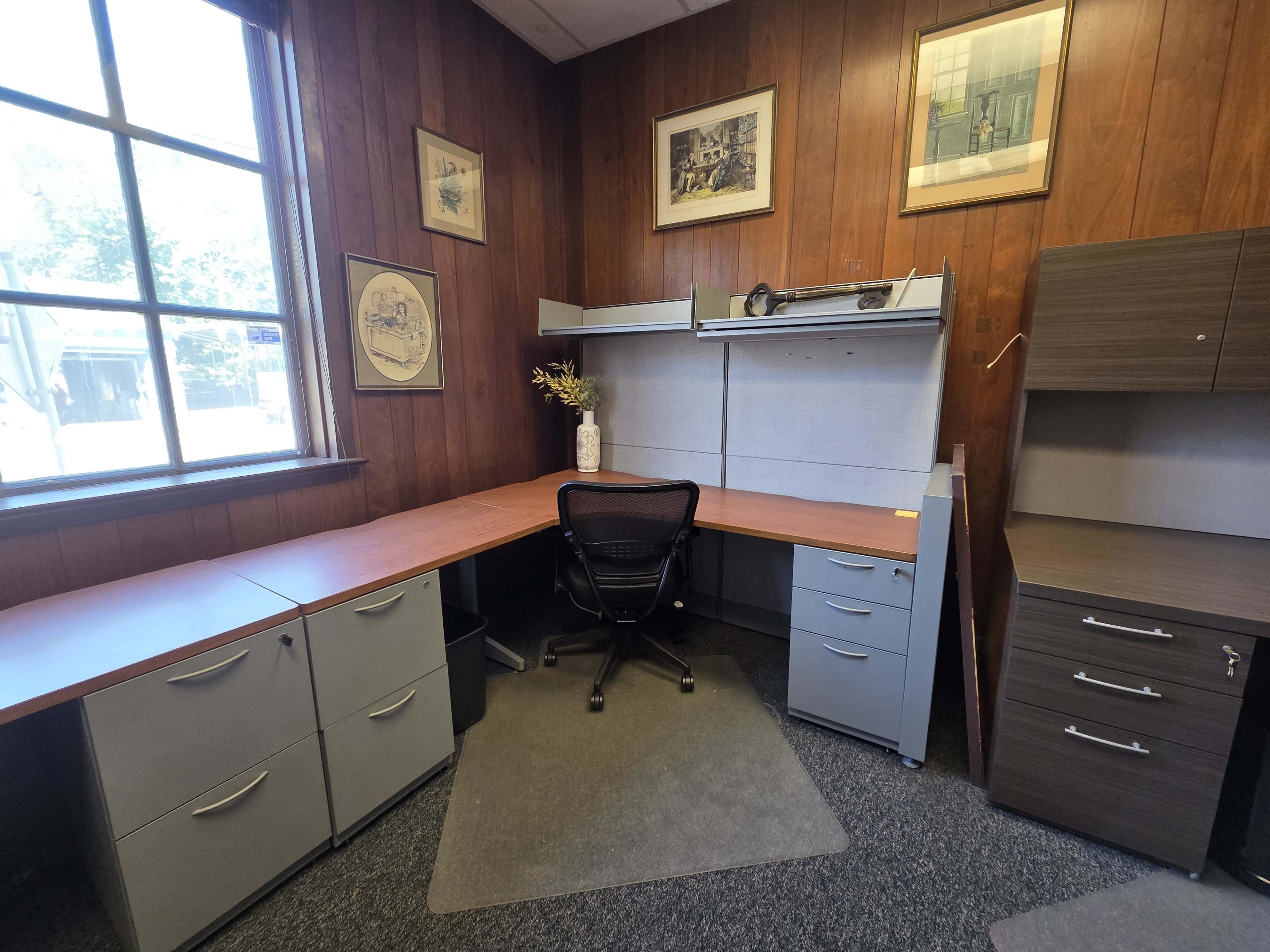 The image shows a small office space with wooden paneling, a desk with storage drawers, an office chair, and a corner shelf.