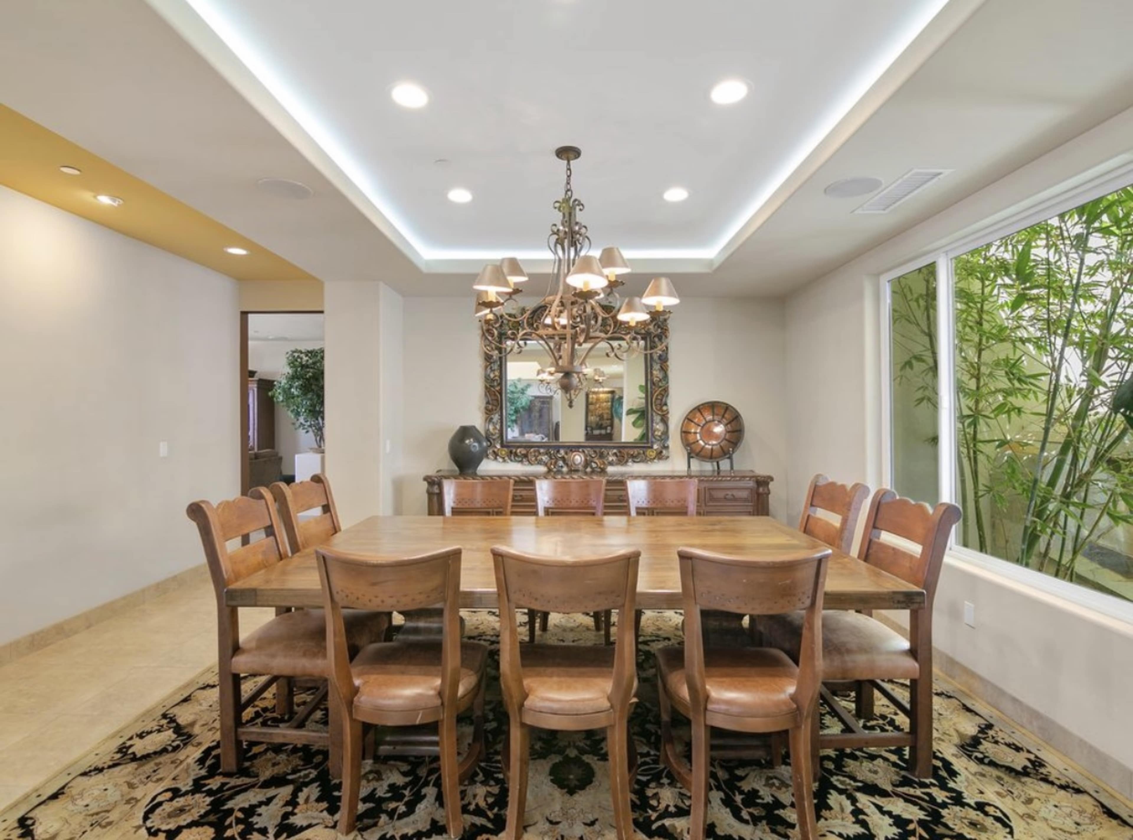 A dining room features a large wooden table surrounded by eight chairs, with a decorative chandelier overhead and a mirror on the wall next to a window showcasing bamboo outside.