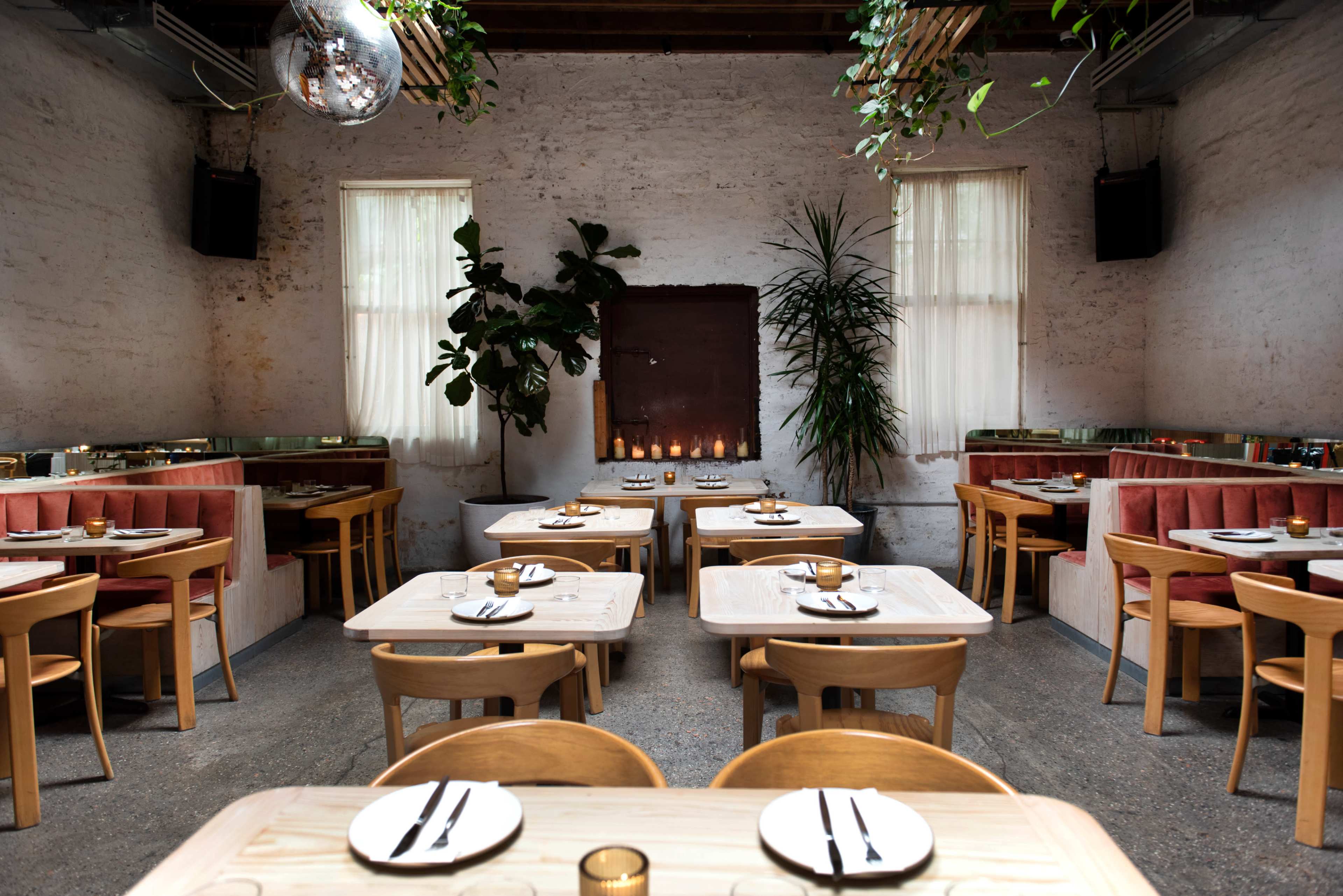 A modern restaurant interior features wooden tables and chairs, with plants and candles adding decor along the walls.