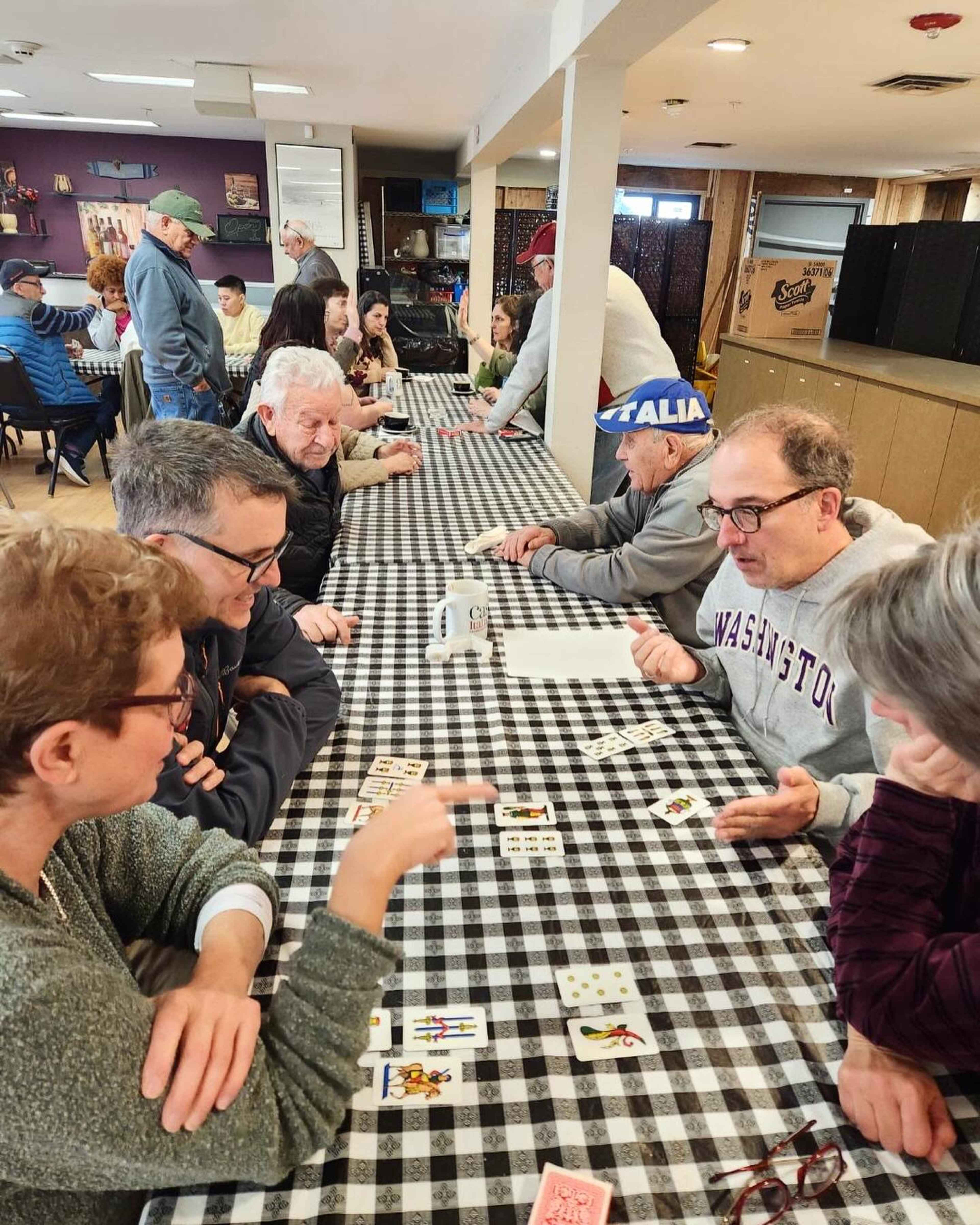 A group of people are seated at a long table covered with a checkered tablecloth, engaged in playing cards and conversing.