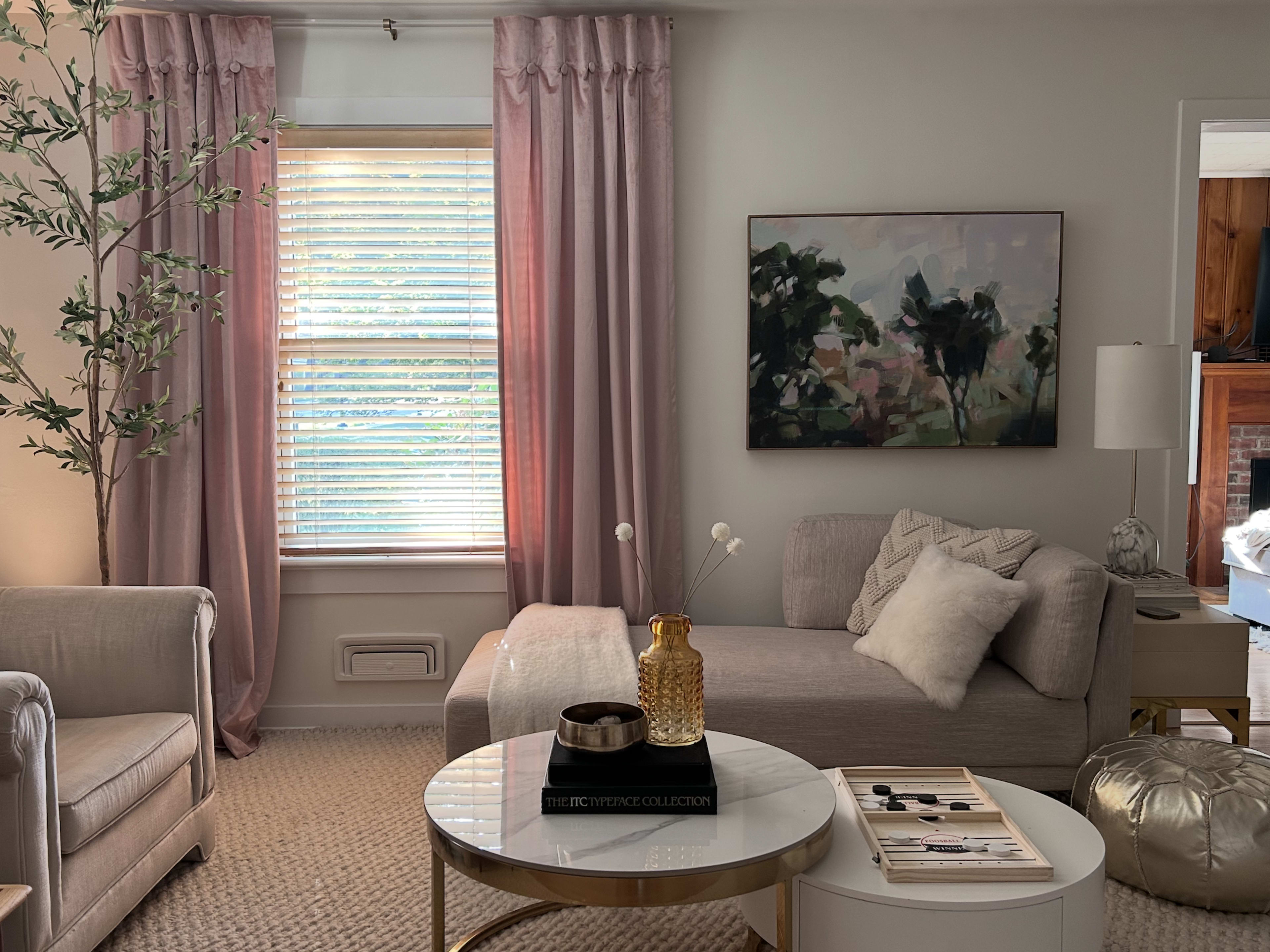 The image shows a bright living room with a light-colored sofa, a round coffee table, and large windows dressed with pink curtains.