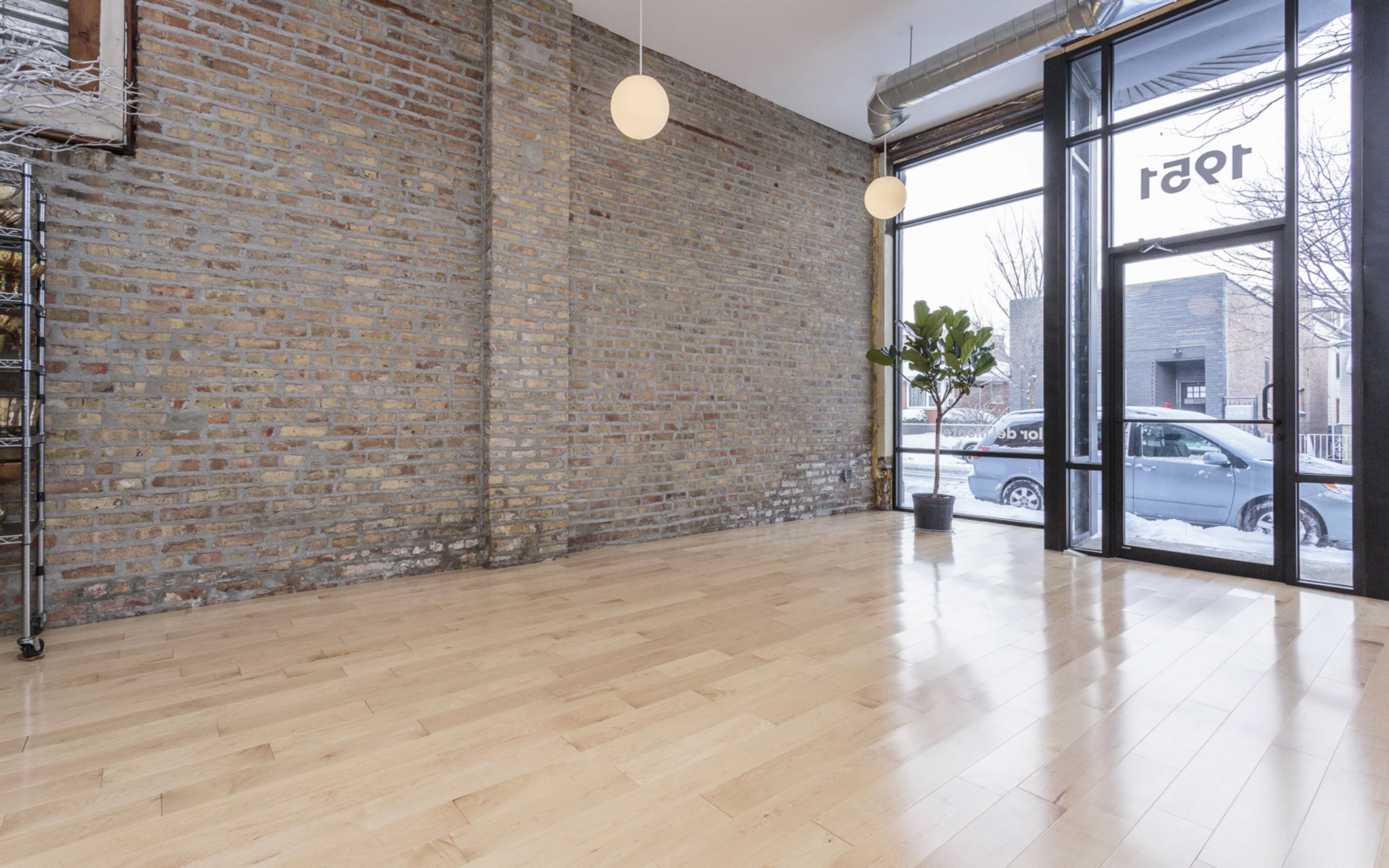 An empty room with wooden floors, large windows featuring a view of a street, and exposed brick walls.