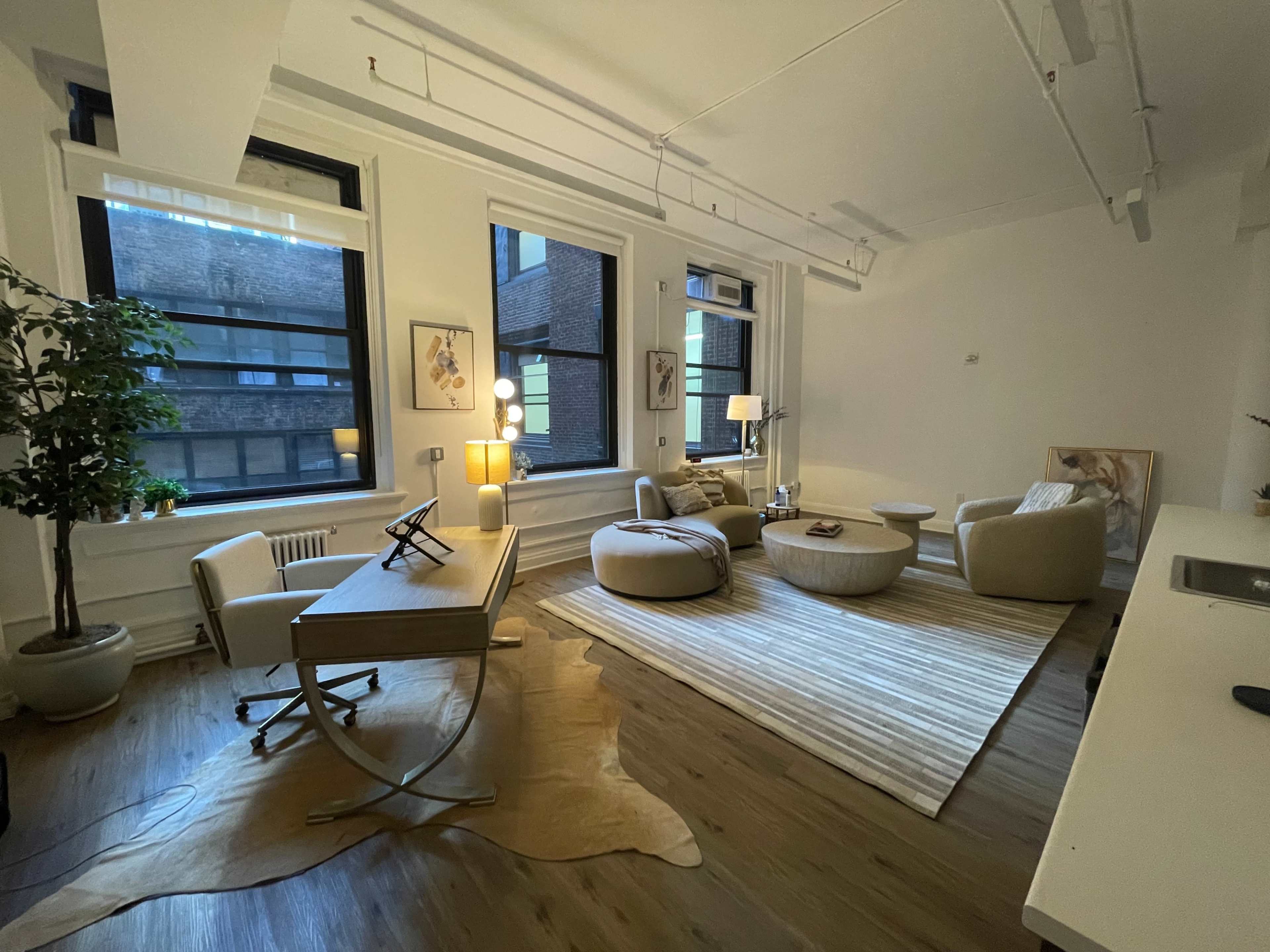 A modern, well-lit office space featuring a desk, a cozy seating area with rounded furniture, and large windows overlooking a cityscape.