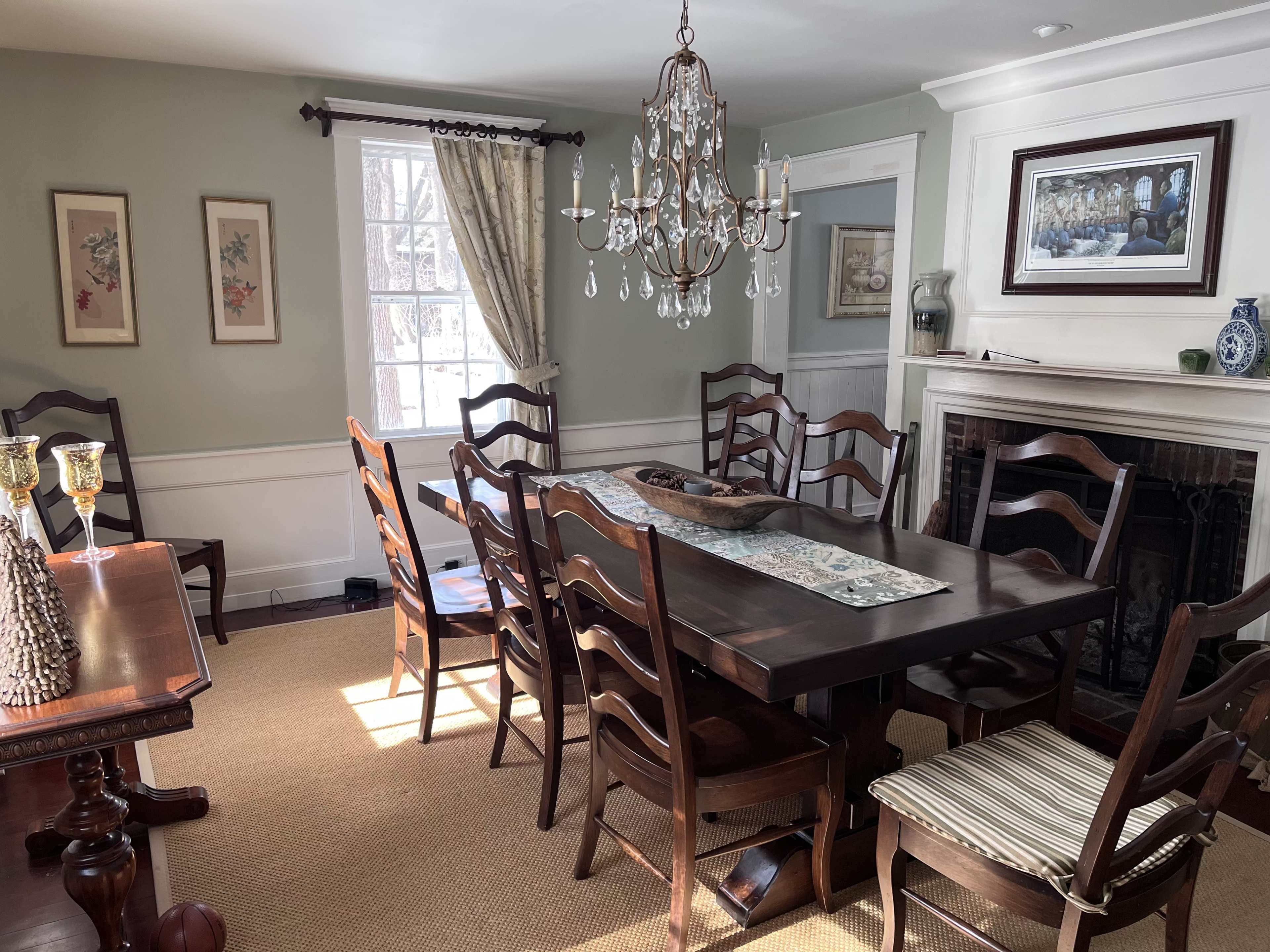 A dining room features a large wooden table surrounded by chairs, with a chandelier overhead and a fireplace against one wall.