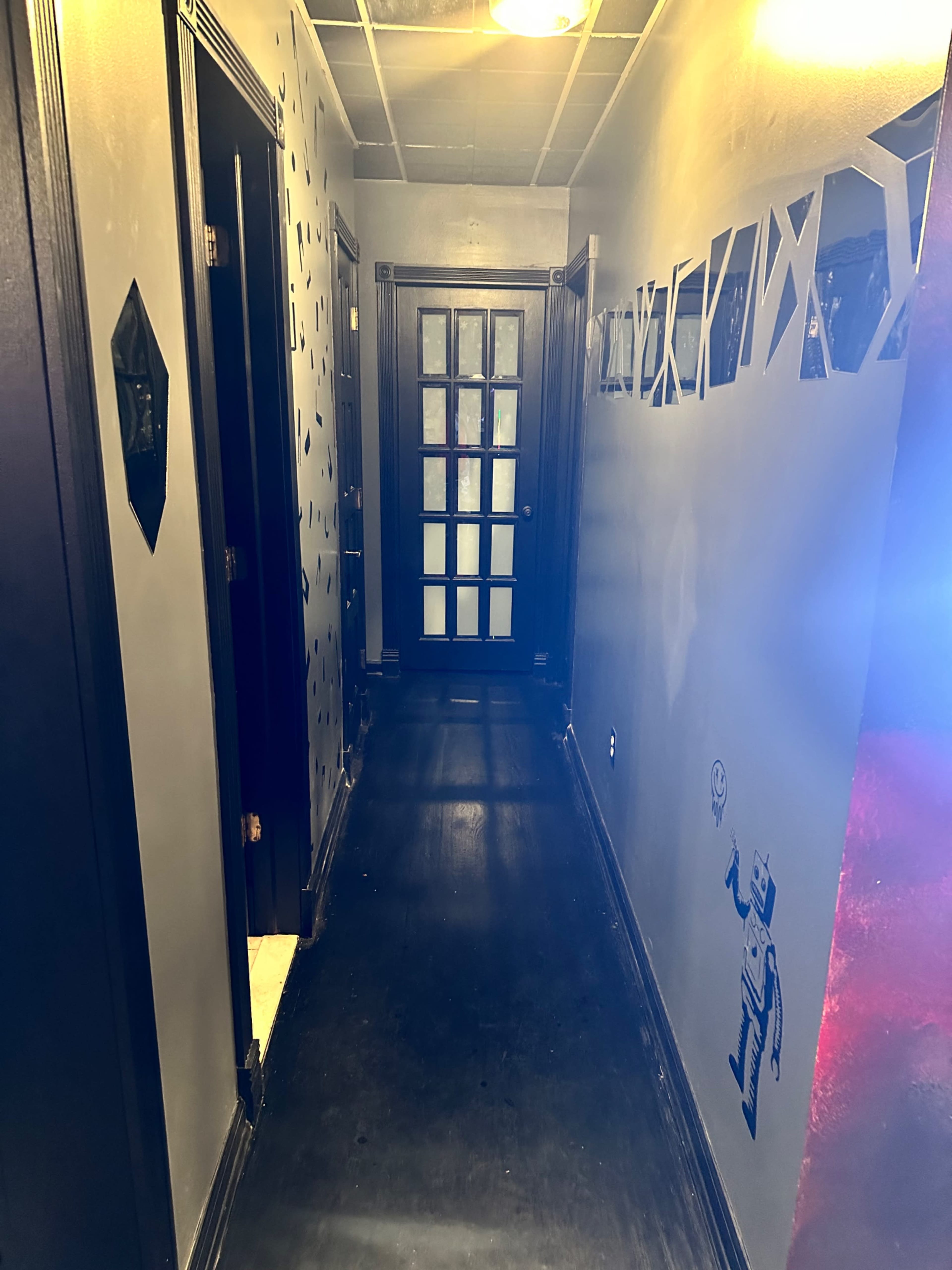 The image shows a narrow hallway with dark walls, geometric decorations, and a door at the far end.