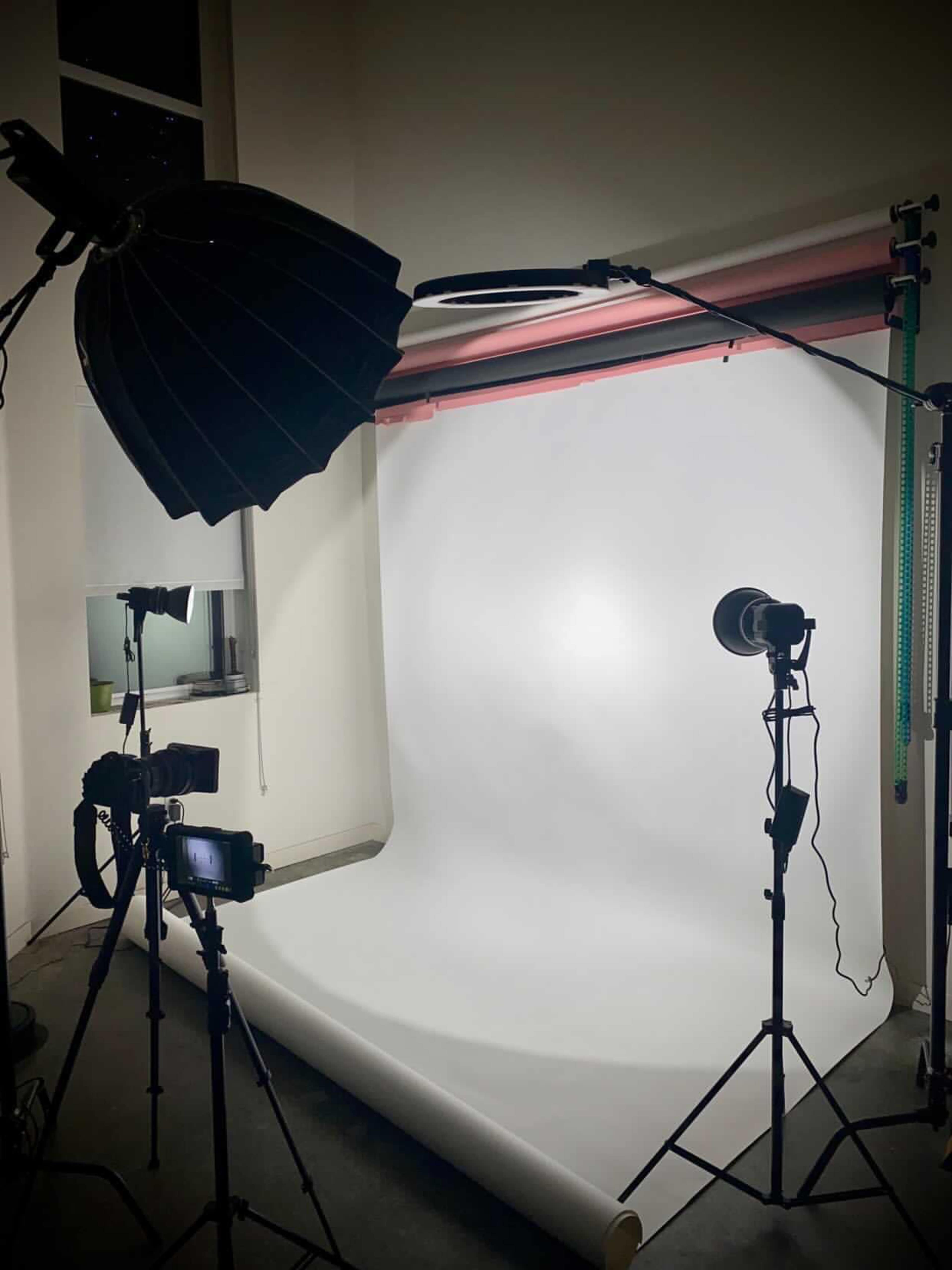 A photography studio setup features a white backdrop, several lighting equipment, and camera tripods.