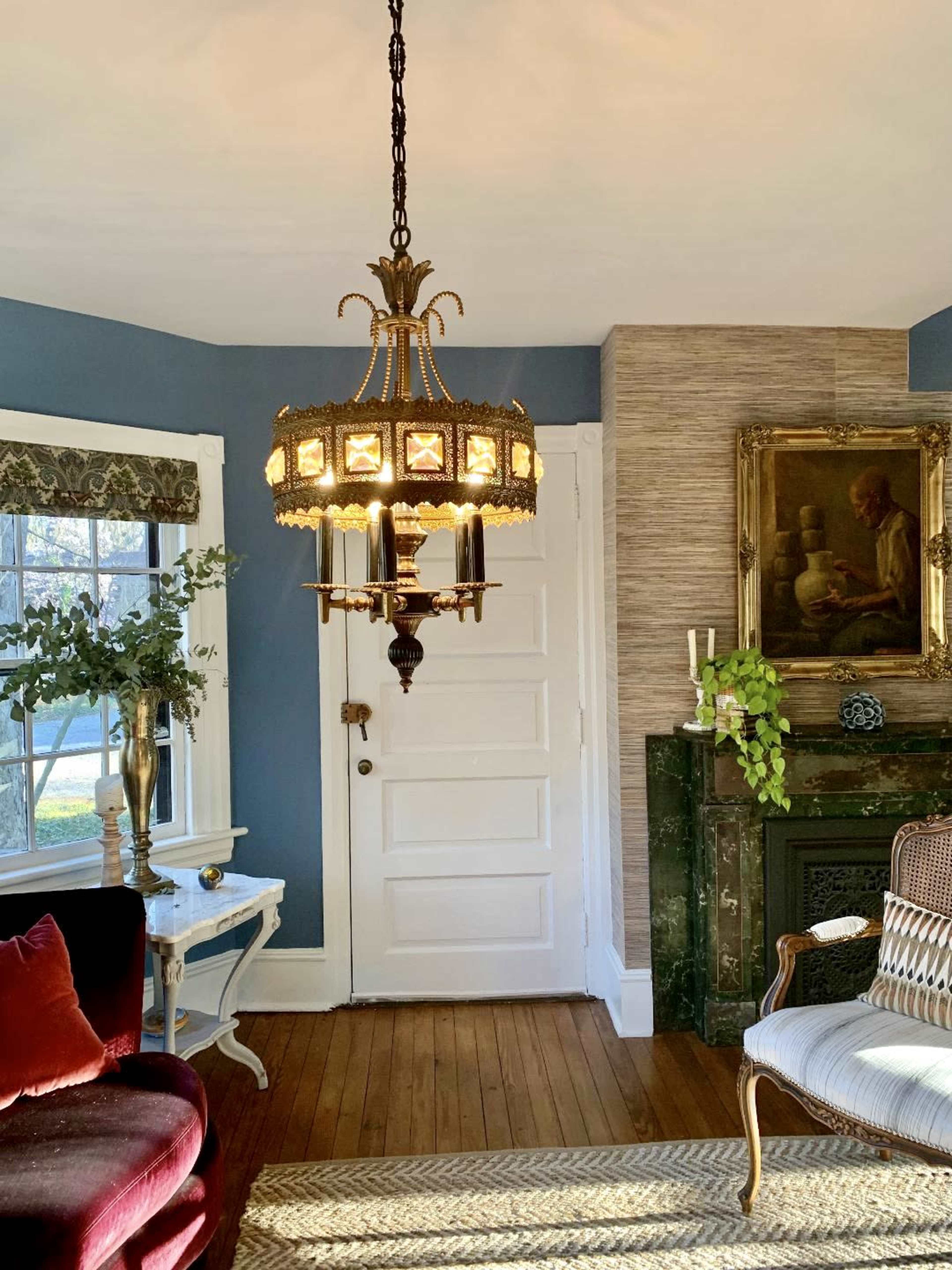 A decorative chandelier hangs from the ceiling in a room with blue walls and hardwood flooring, featuring a white door and a fireplace with a painting above it.