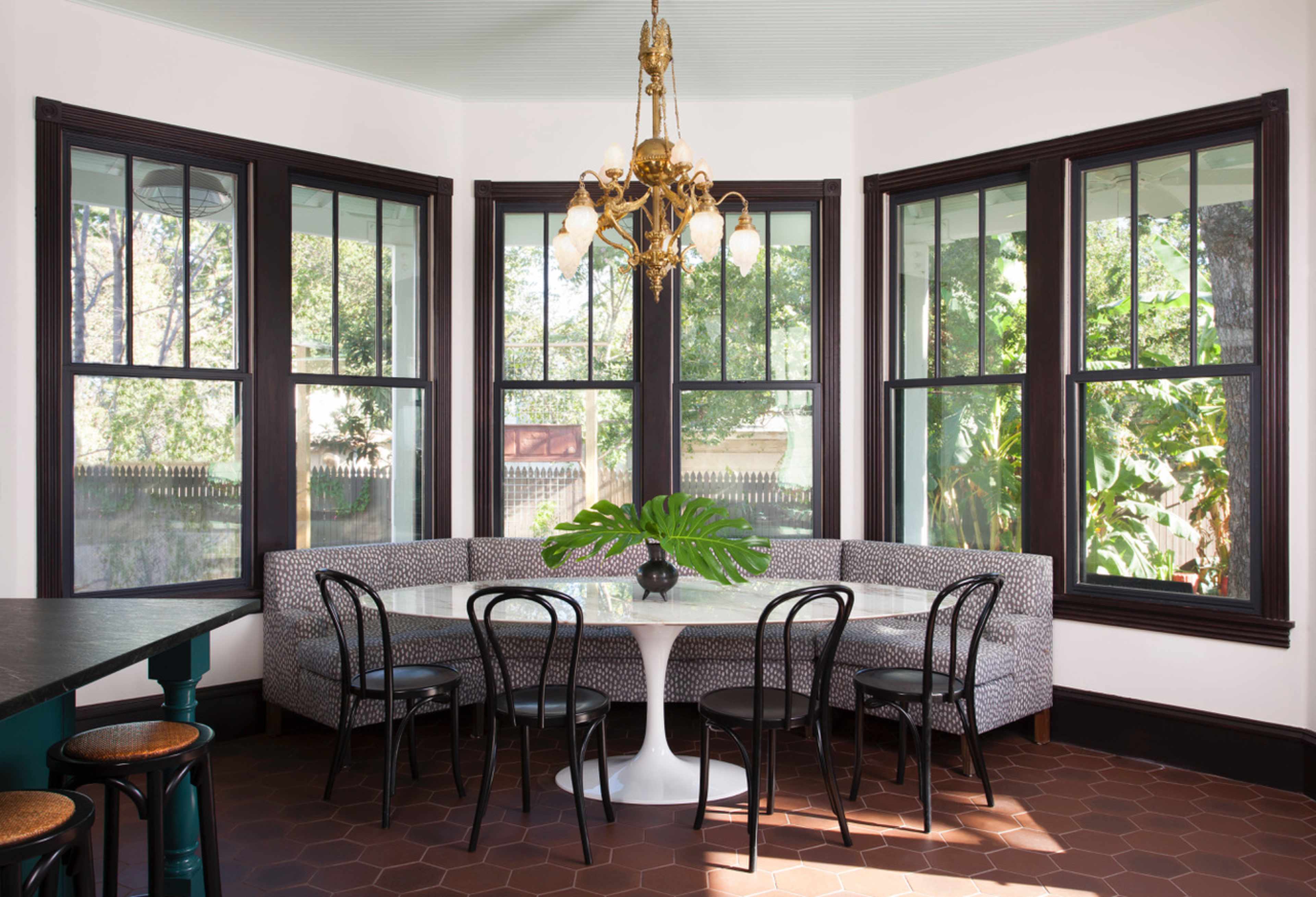 The image shows a dining area featuring a round white table surrounded by black chairs, with a curved gray banquette and large windows providing a view of the outdoors.