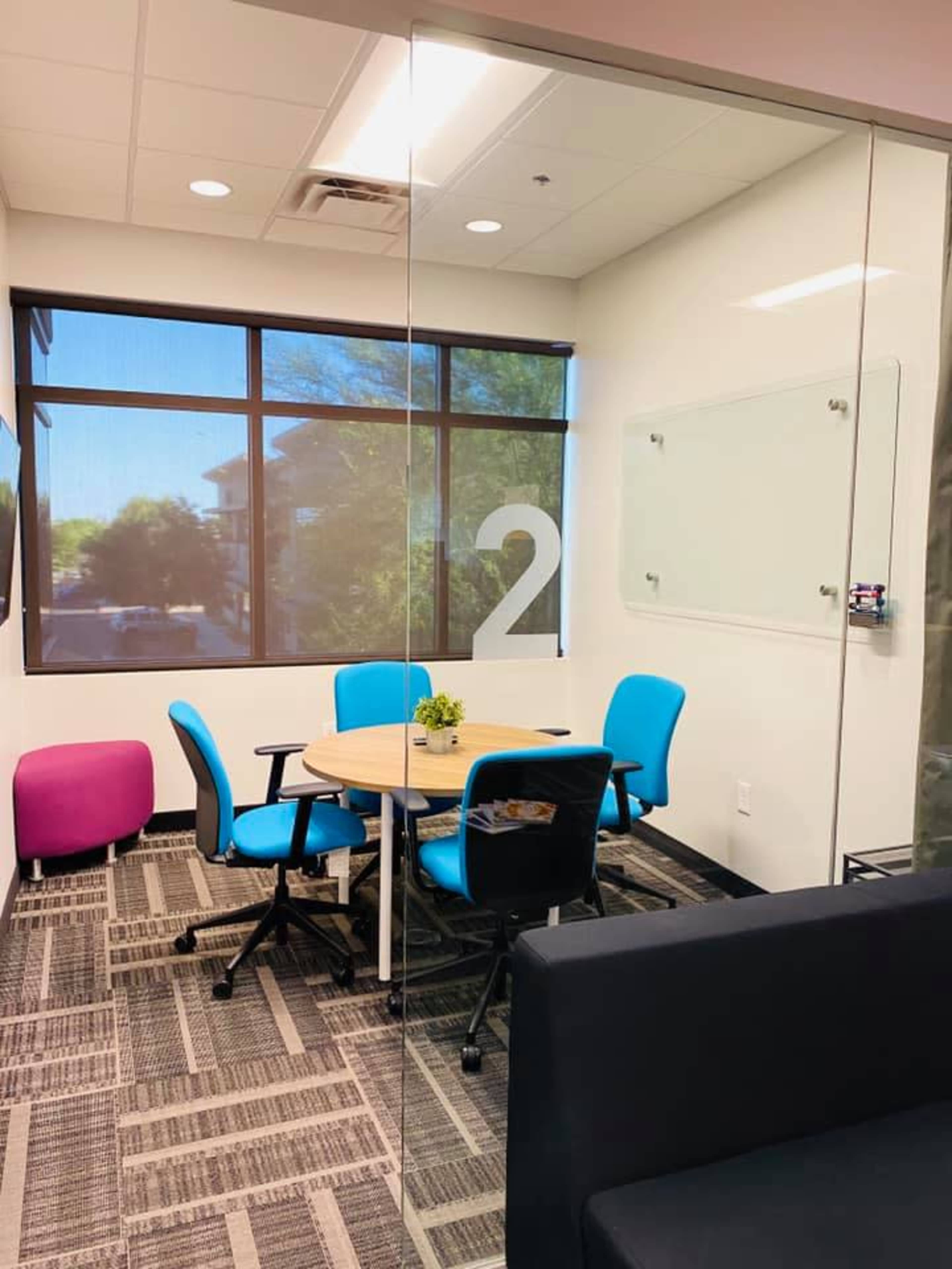 The image shows a small meeting room with a round table and four blue chairs, featuring a large glass wall displaying the number "2."