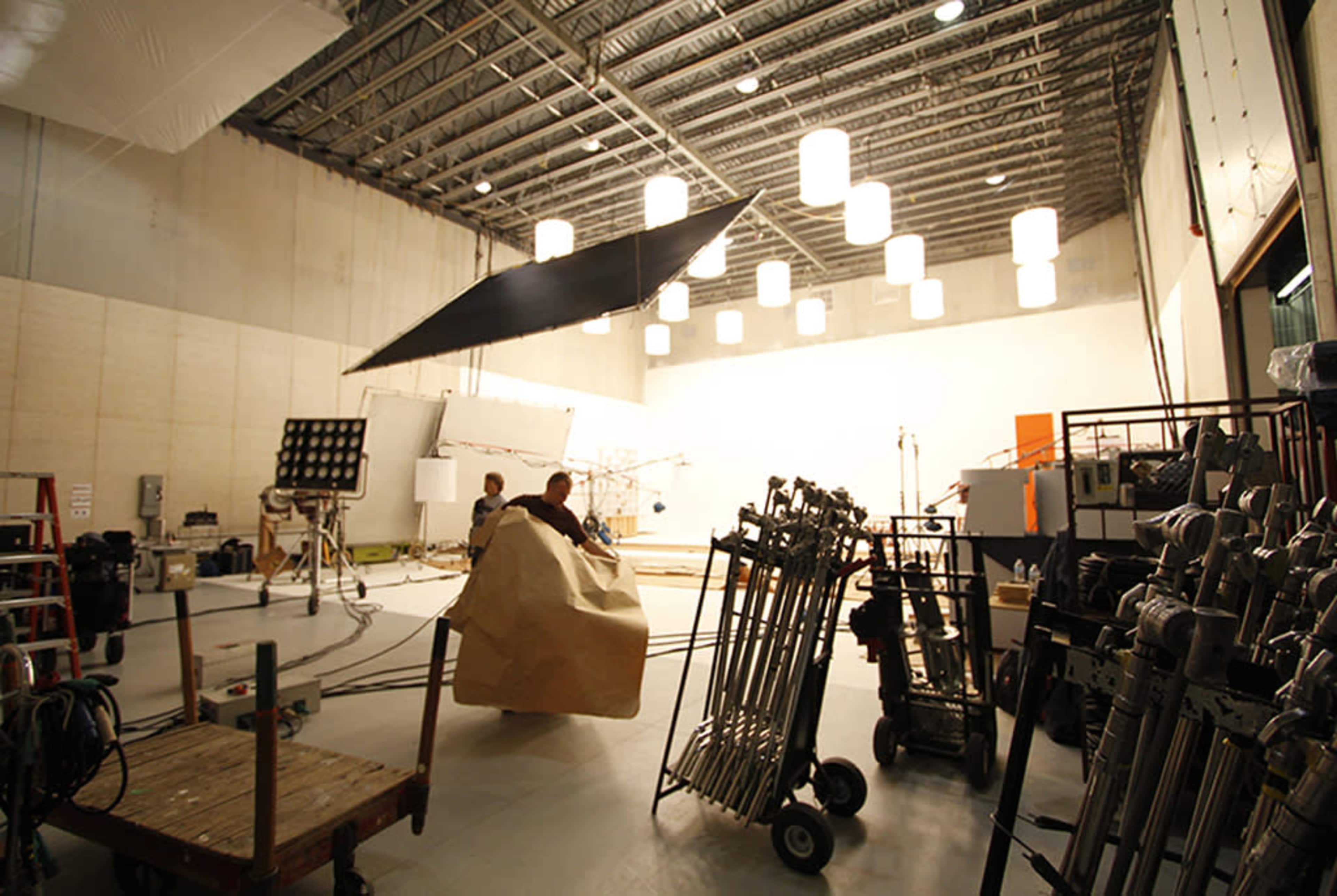 A film set is shown with crew members preparing equipment under overhead lighting in a spacious studio.