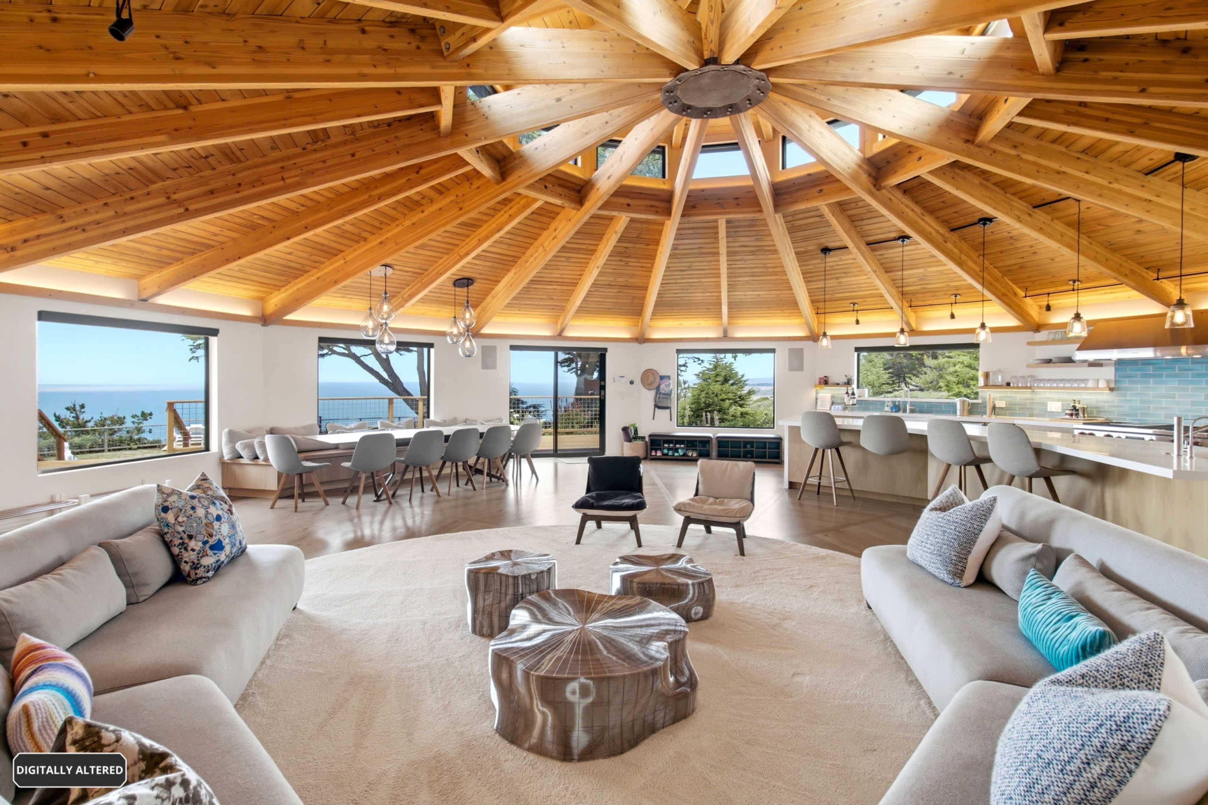 The image shows a modern, circular living space with a wooden, vaulted ceiling, large windows offering ocean views, and a mix of couches and tables arranged around a central area.