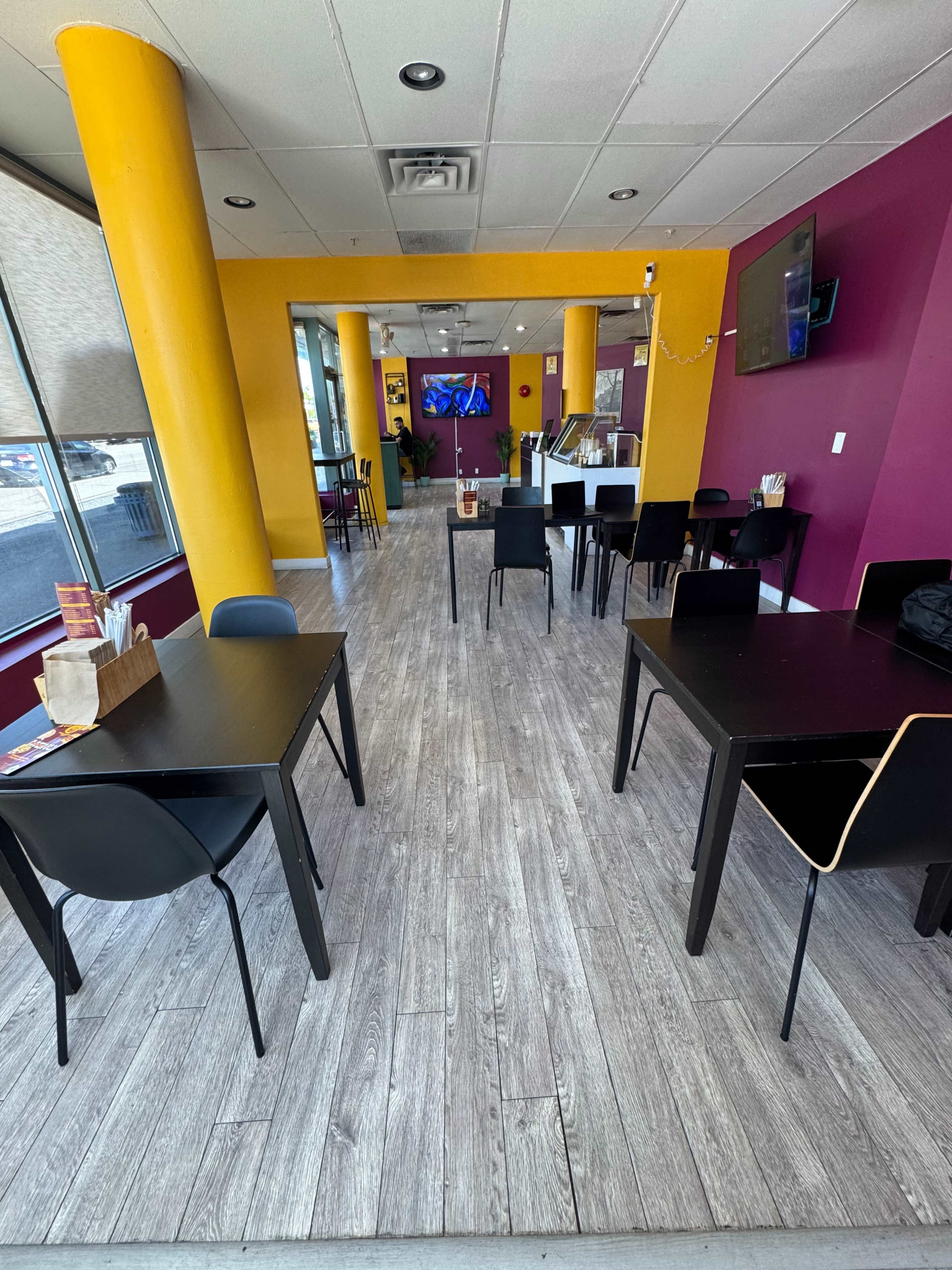The image shows a modern café interior featuring black tables and chairs, colorful purple and yellow walls, and a large television on the wall.