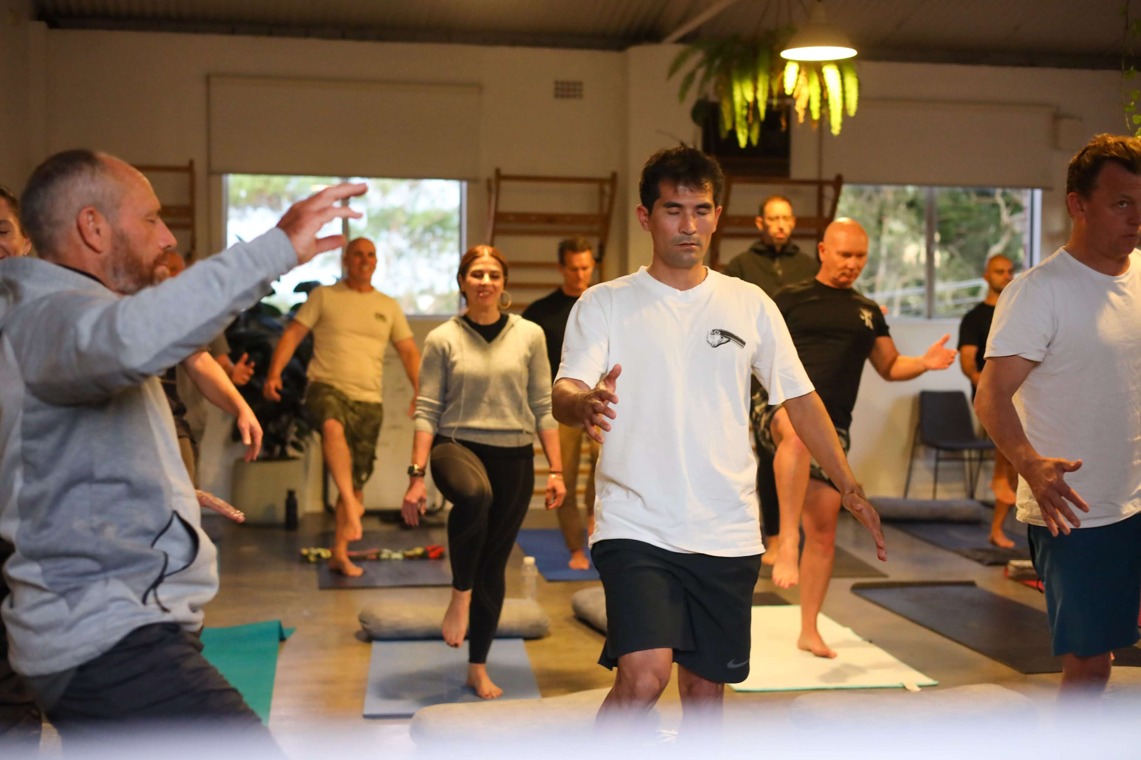 A group of people practice yoga in a spacious studio with mats and cushions on the floor.