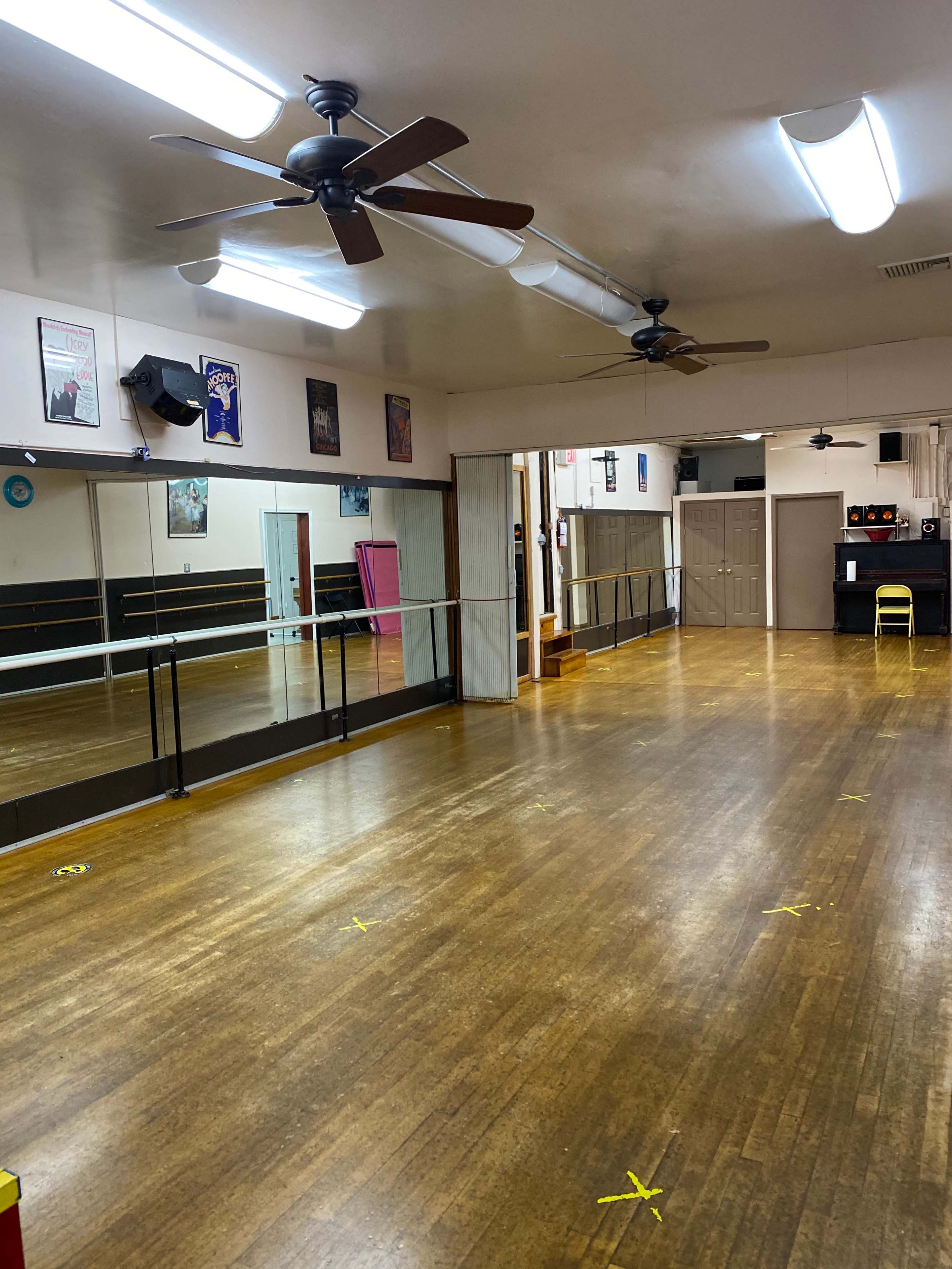 A large dance studio features wooden floors, wall mirrors, and ceiling fans, with marked spots on the floor for spacing.