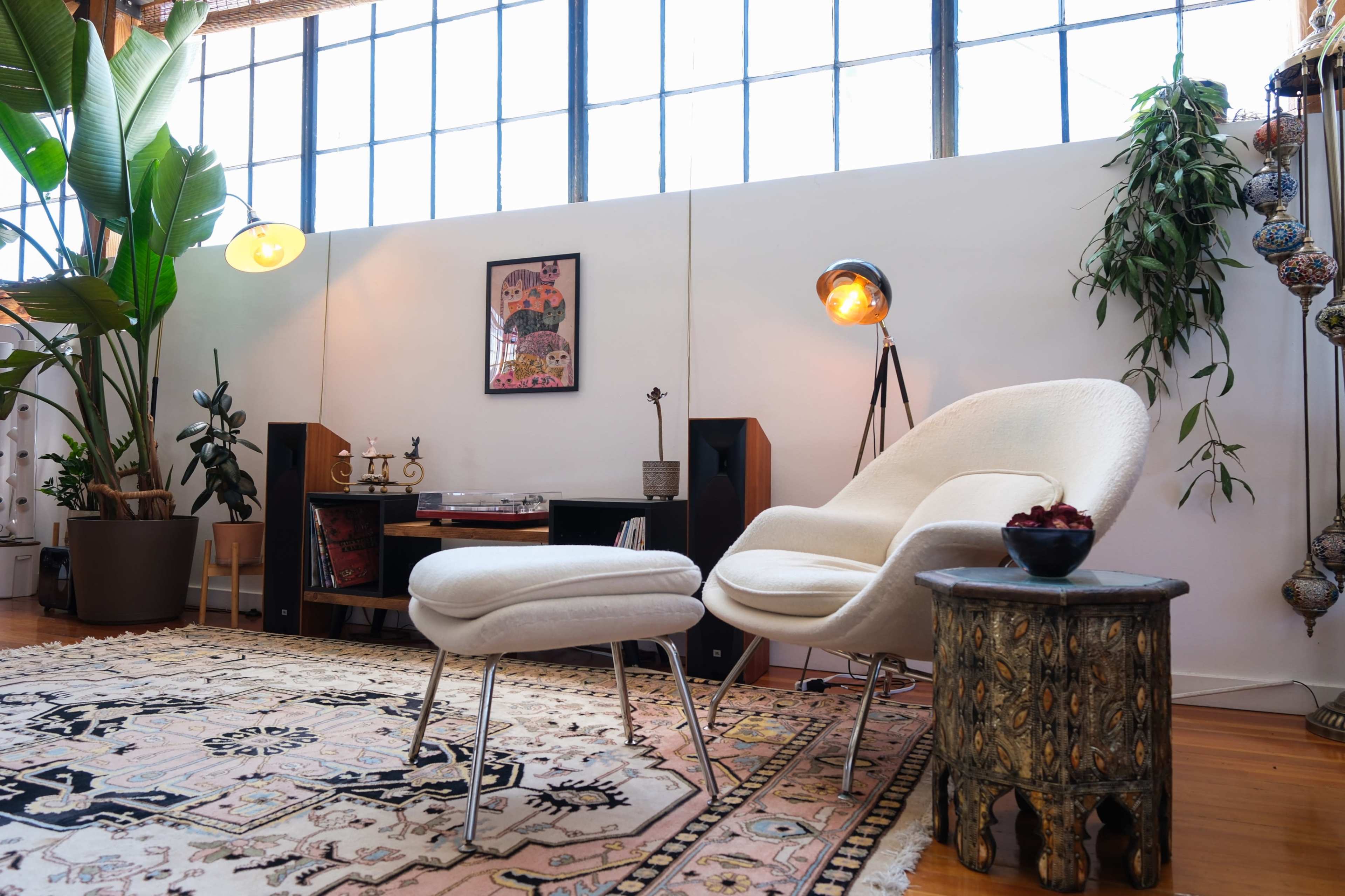 The image shows a modern living room featuring a white armchair and ottoman paired with a wooden side table, adorned with a decorative rug and large windows letting in natural light.