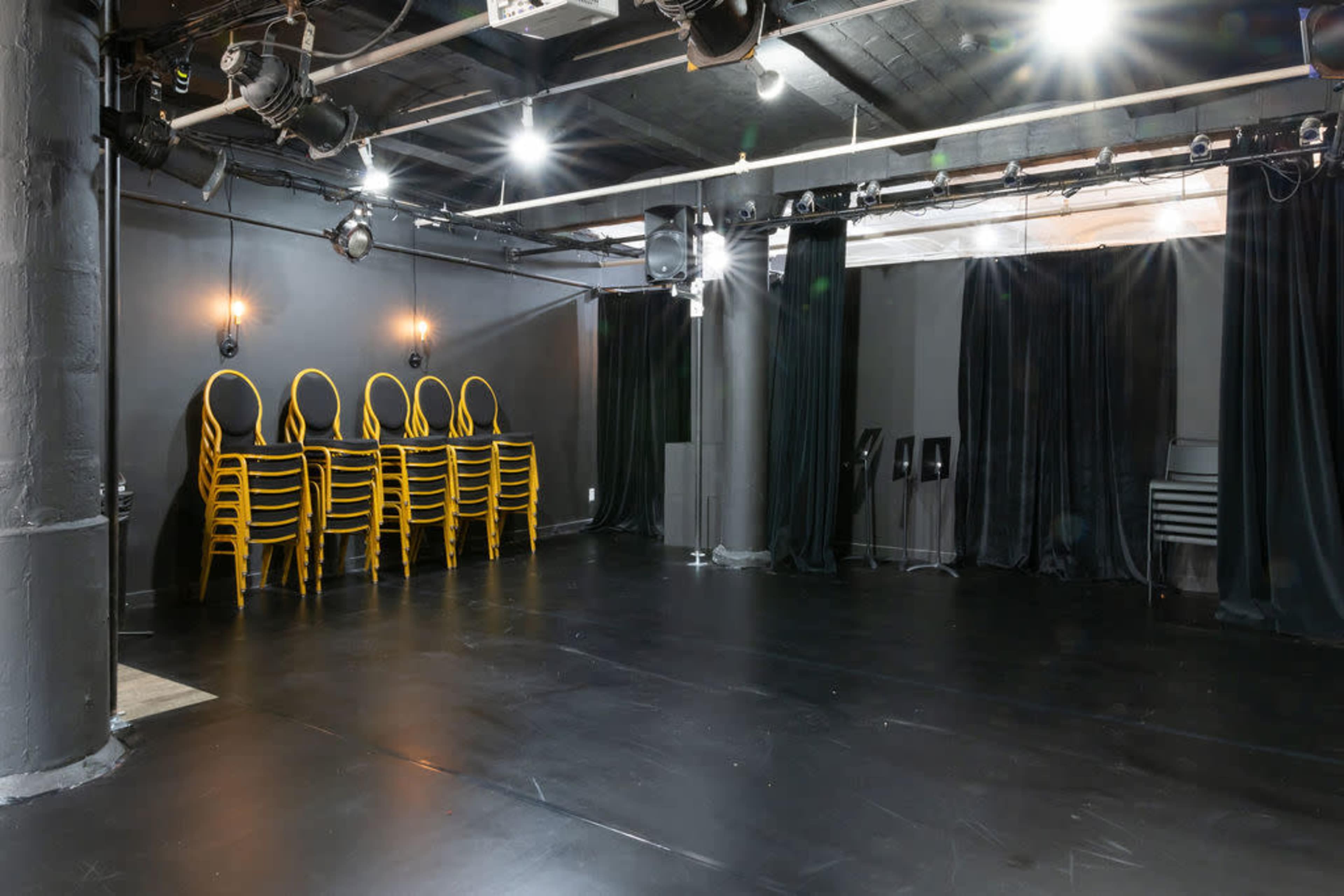 A large, empty performance space with black flooring, stacked yellow chairs, and curtains along the stage area.