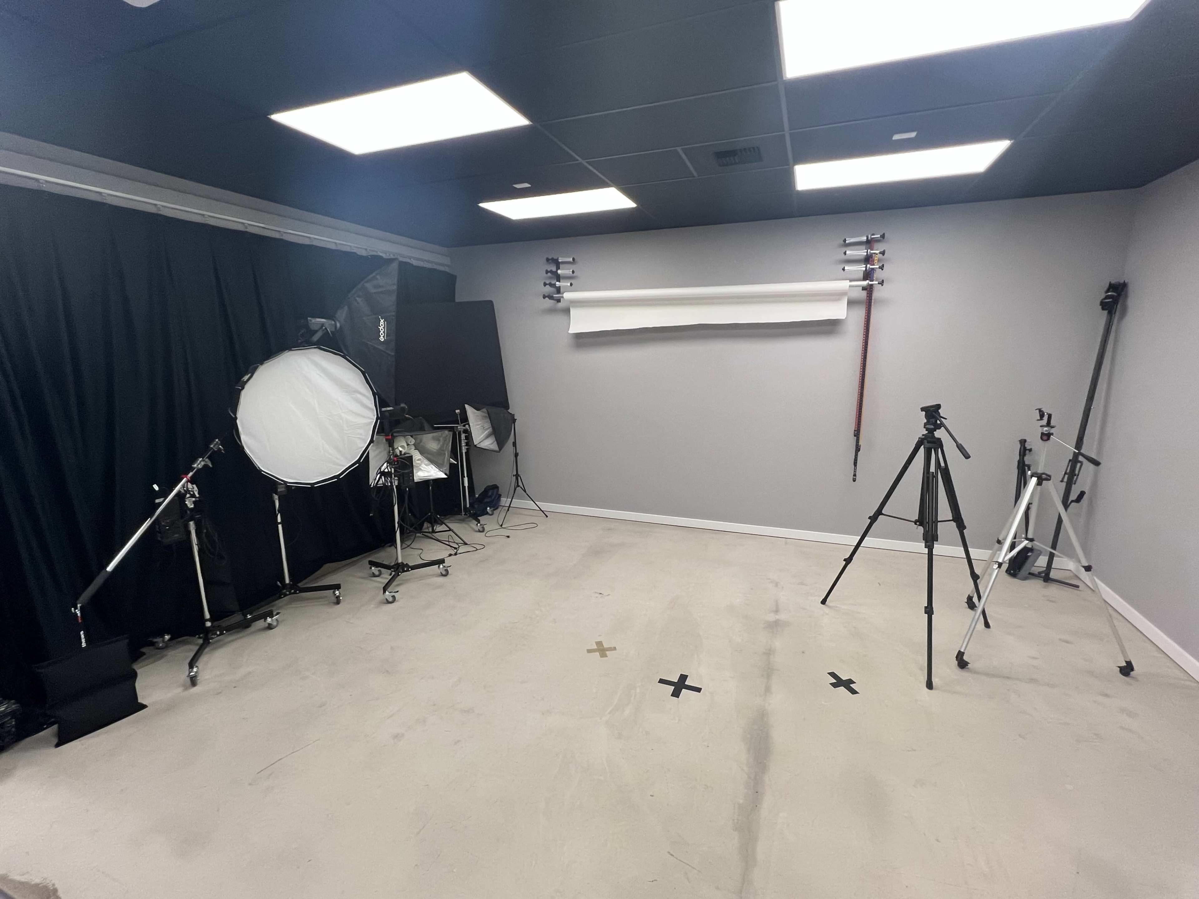 The image shows a photography studio with various lighting equipment and tripods arranged against a gray backdrop.