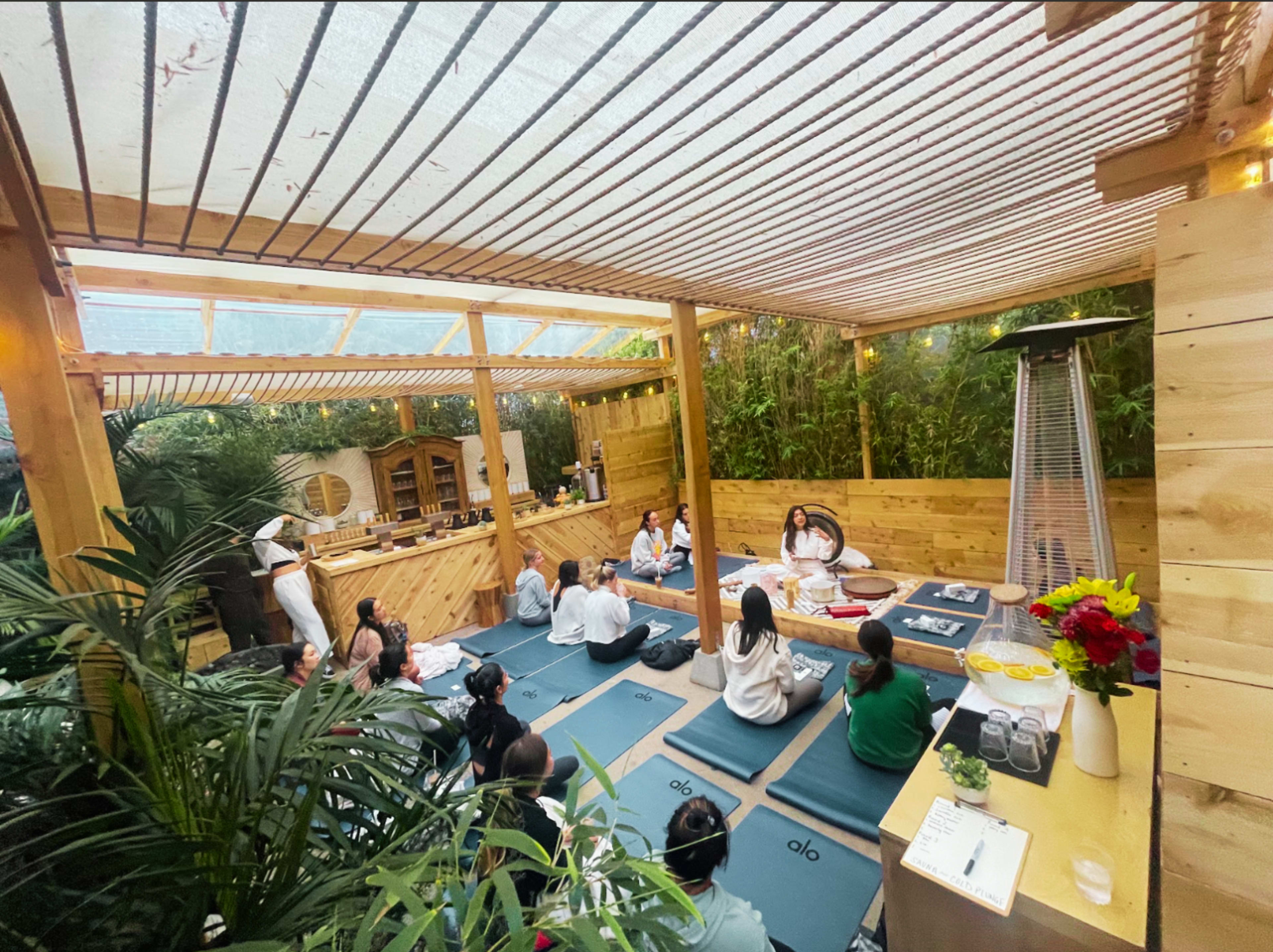 A group of people sit on mats in a wooden outdoor space that features greenery and a bar area.