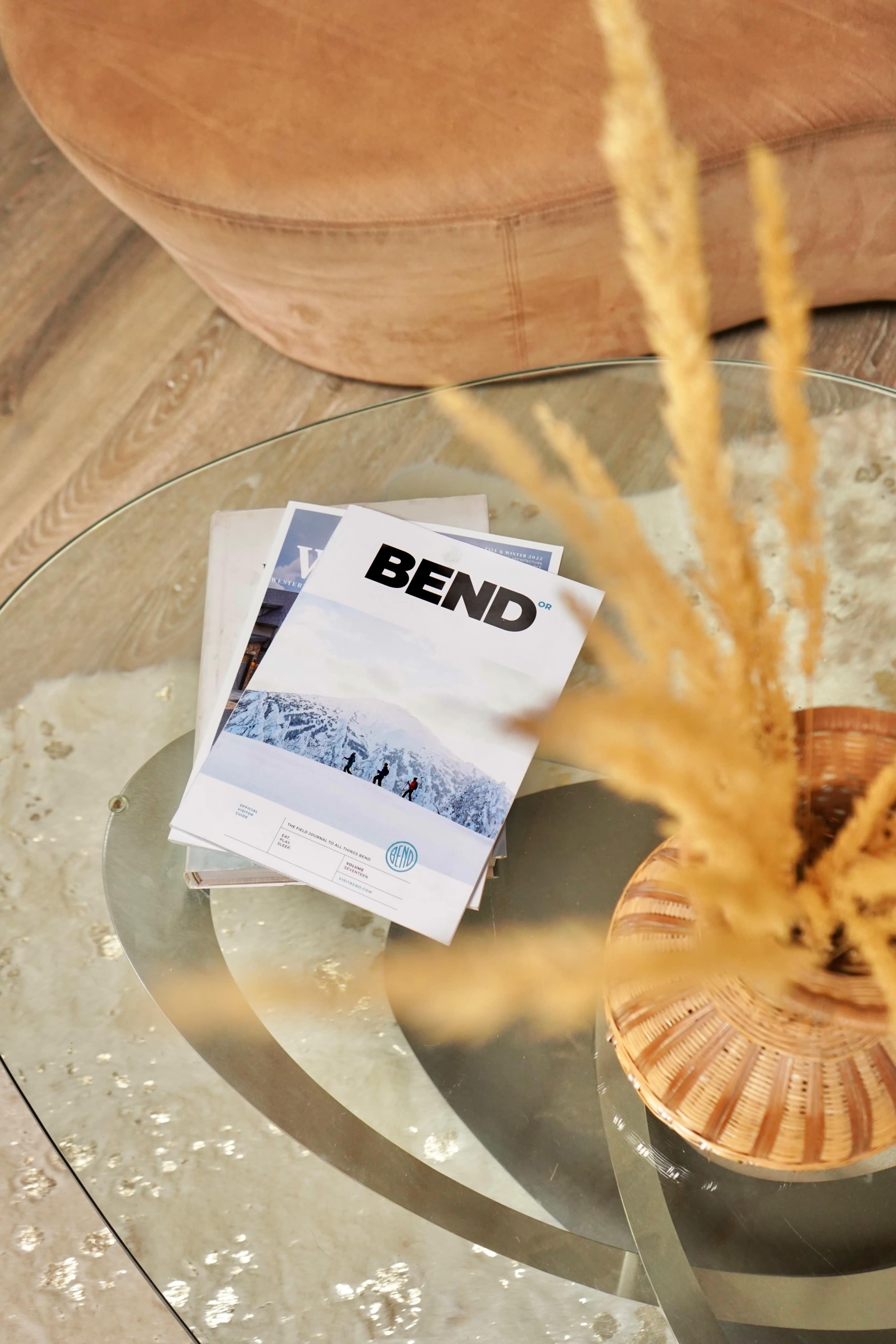 A glass table holds magazines, including one titled "BEND," alongside a decorative vase with dried flowers.