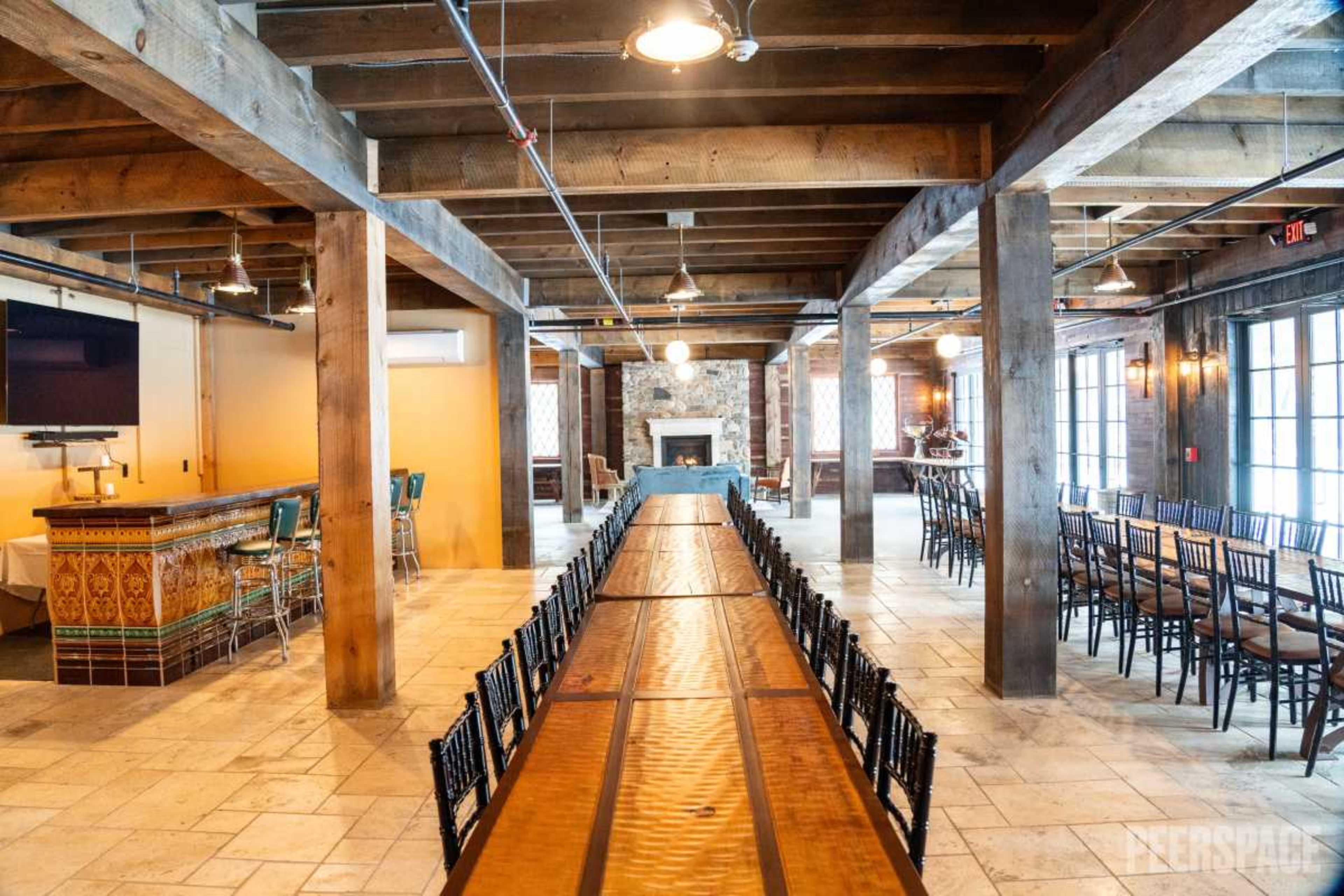 The image shows an indoor venue with exposed wooden beams, featuring a long wooden table and multiple rows of black chairs, alongside a bar area and a fireplace in the background.