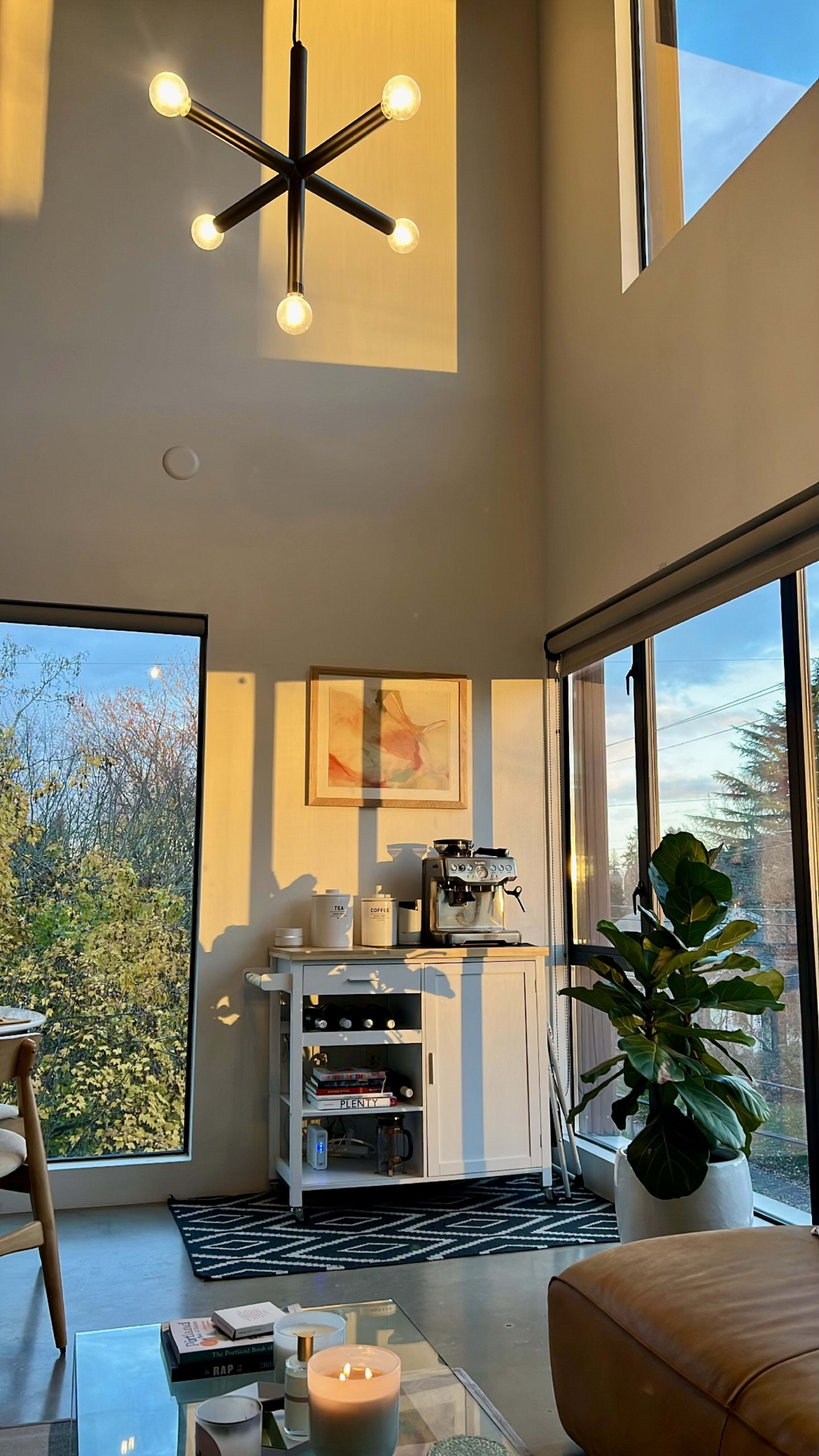 A modern living space with large windows, a coffee station, a potted plant, and a contemporary light fixture.