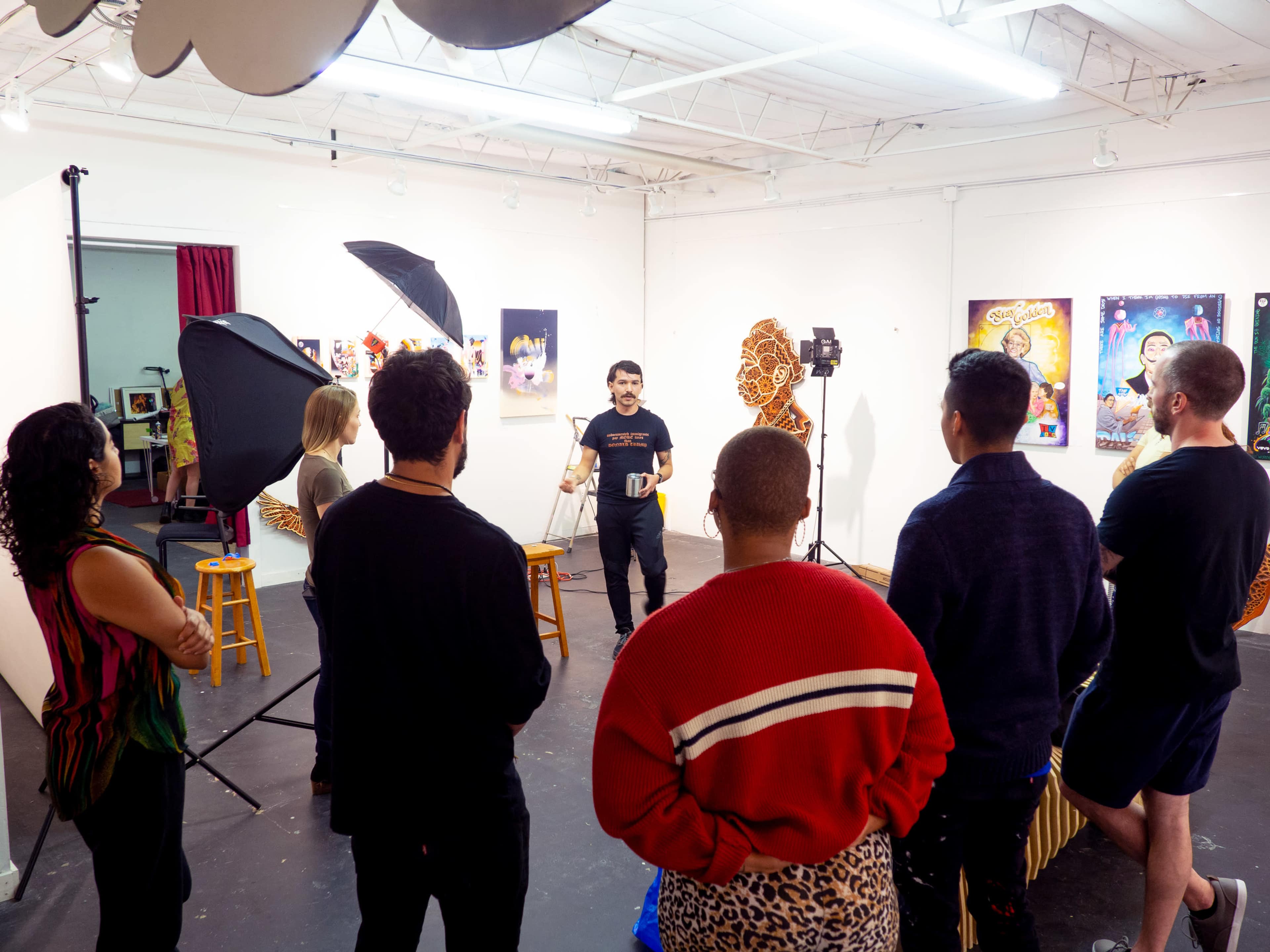 A group of people stands in a circle around a speaker in an art studio, with various lighting equipment and artworks on the walls.