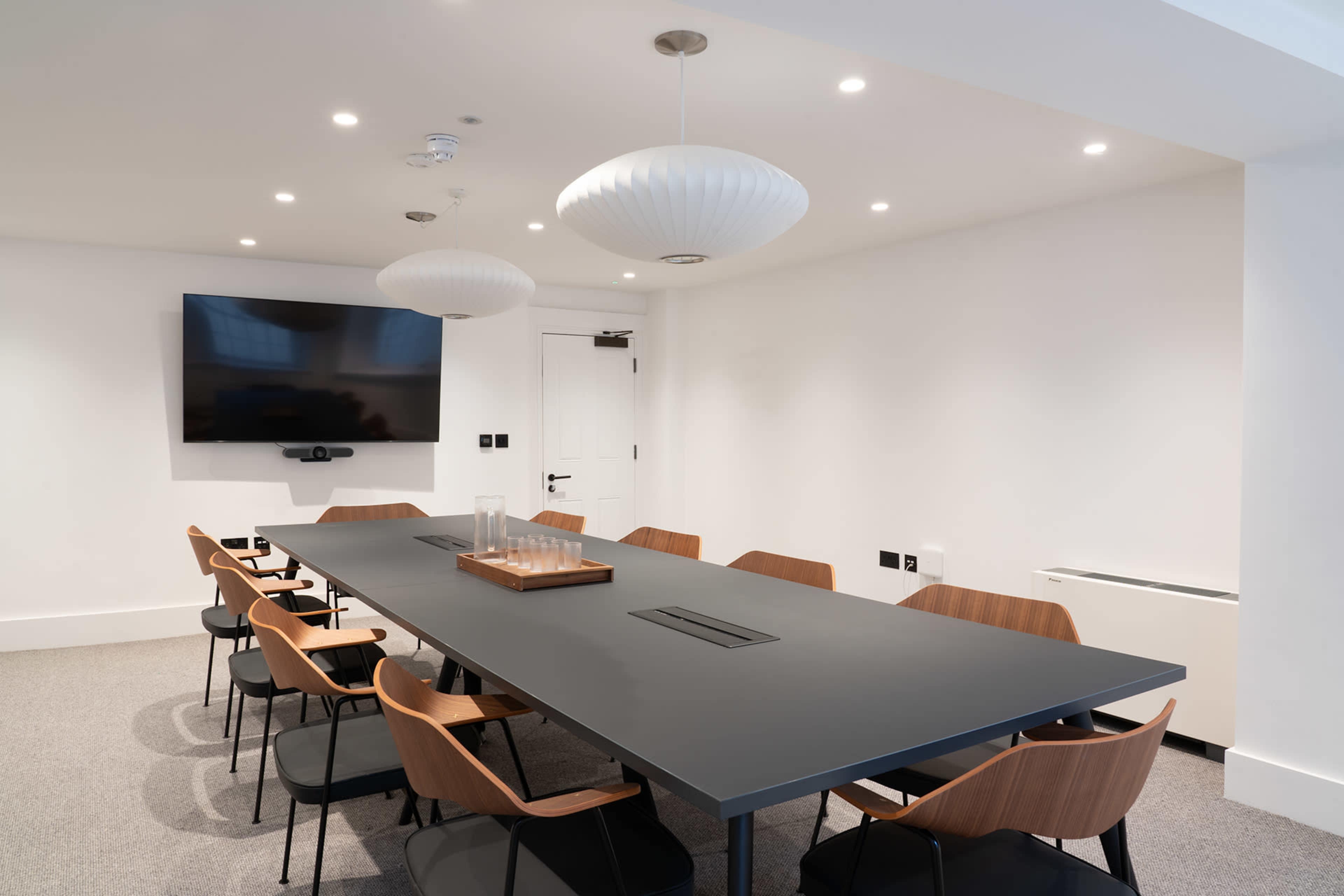 A modern conference room features a long table surrounded by chairs, with a wall-mounted television and two pendant lights above.