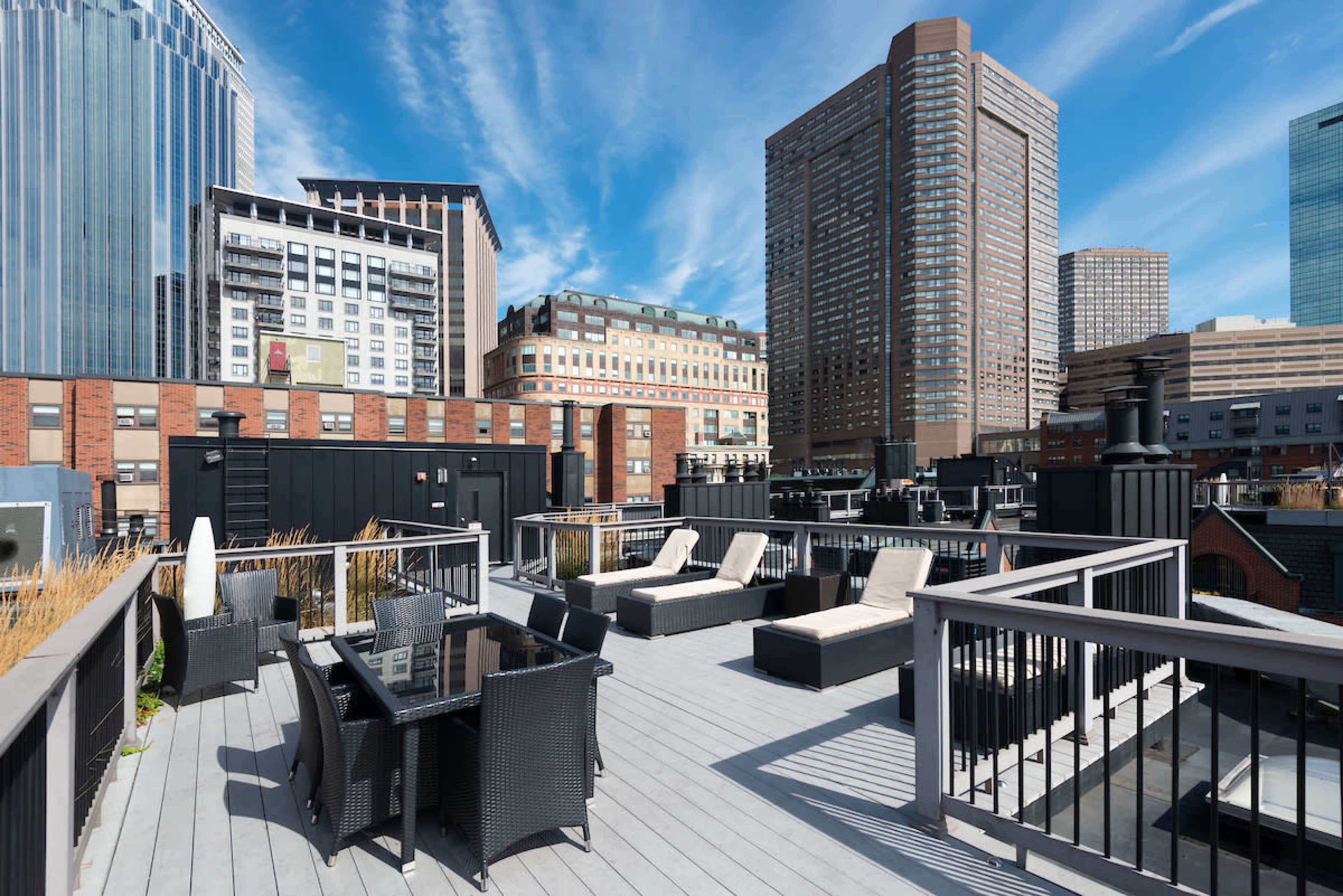 A rooftop terrace with lounge chairs and a dining area, surrounded by tall modern buildings against a clear blue sky.