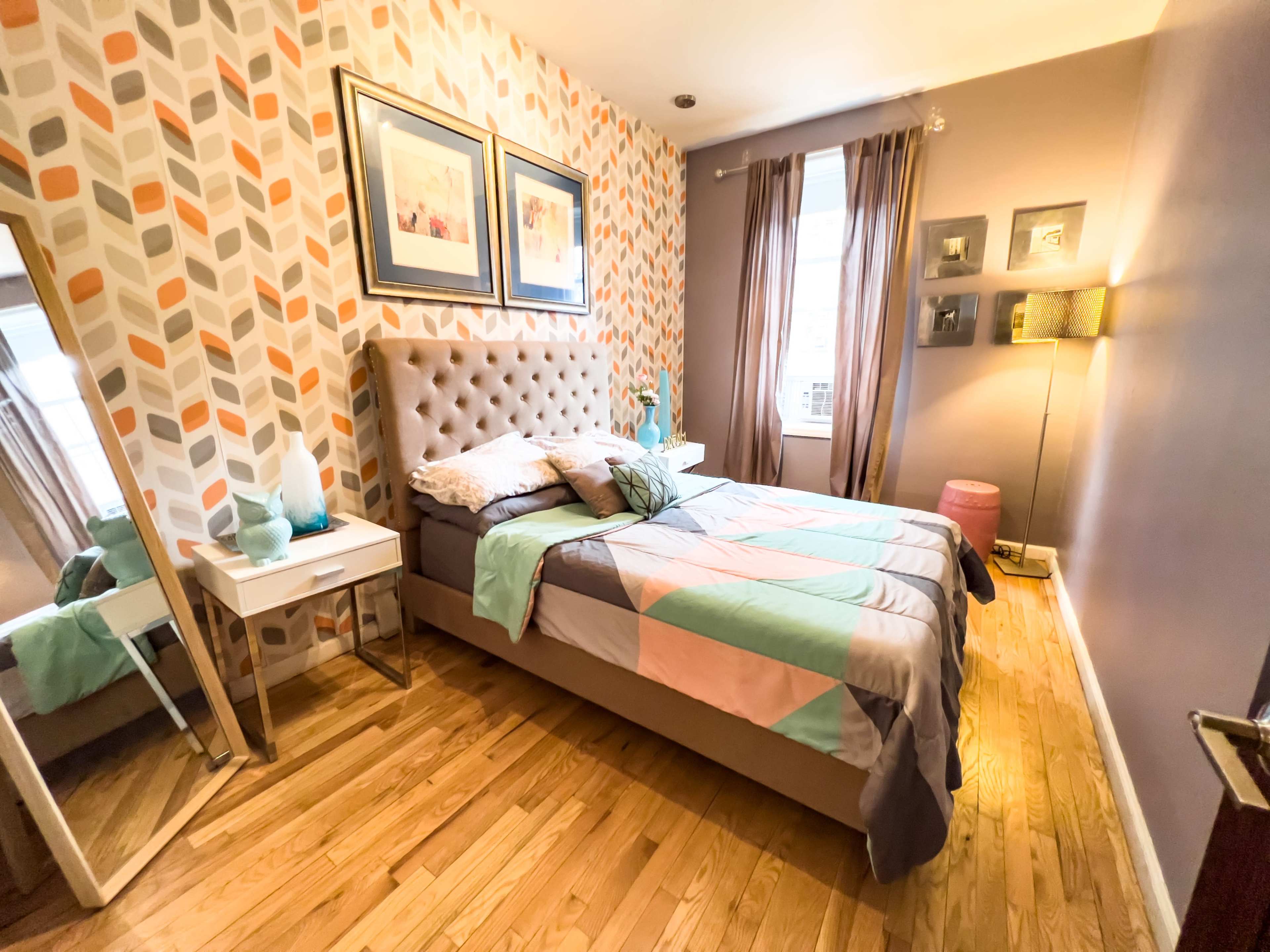 A cozy bedroom with a patterned accent wall, a queen-sized bed, a full-length mirror, and a lamp beside a small side table.