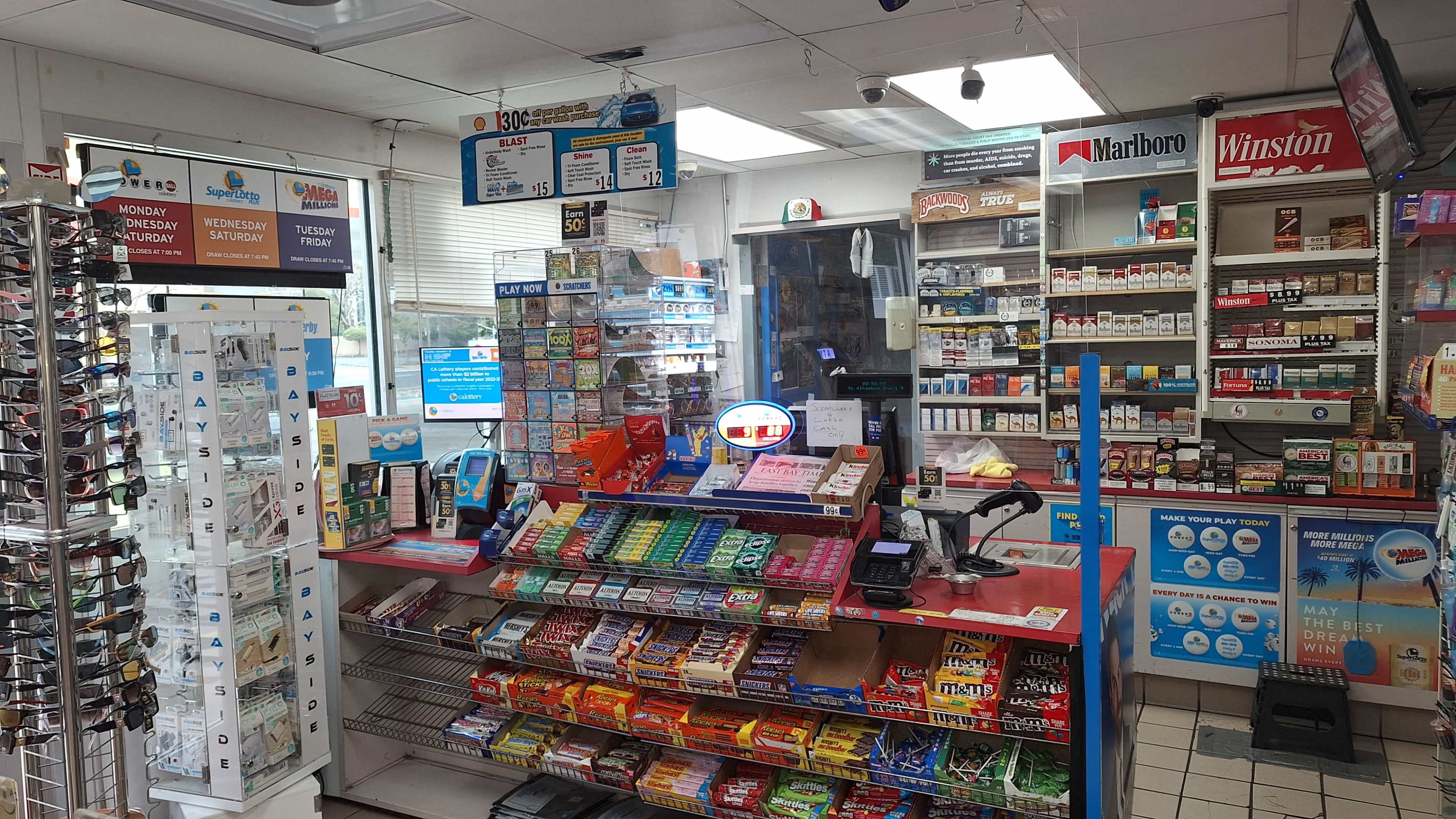 The image shows the interior of a convenience store with shelves stocked with various snacks, beverages, and cigarette brands.