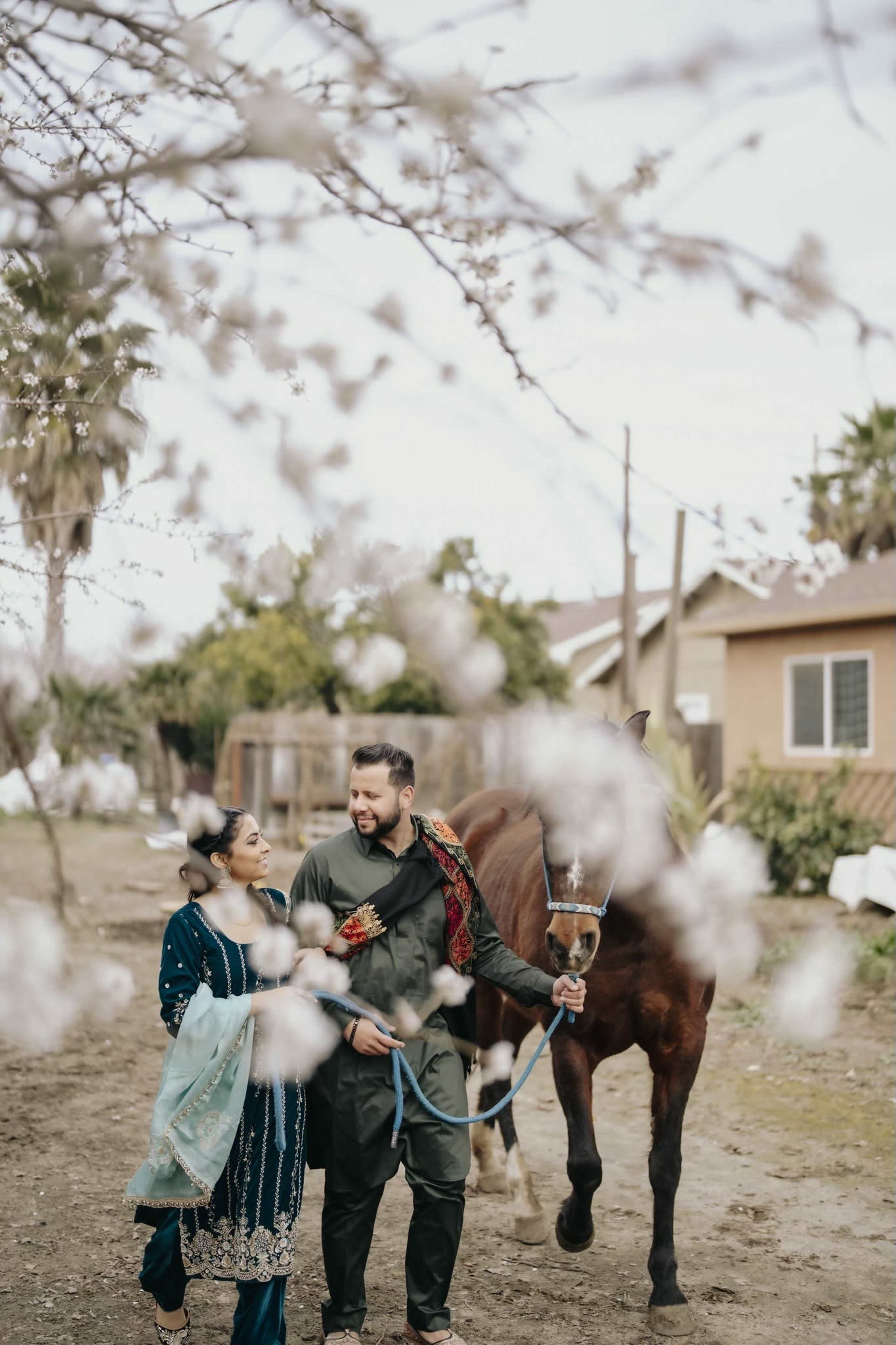 A man and a woman walk together holding a horse near residential buildings, surrounded by blooming flowers.