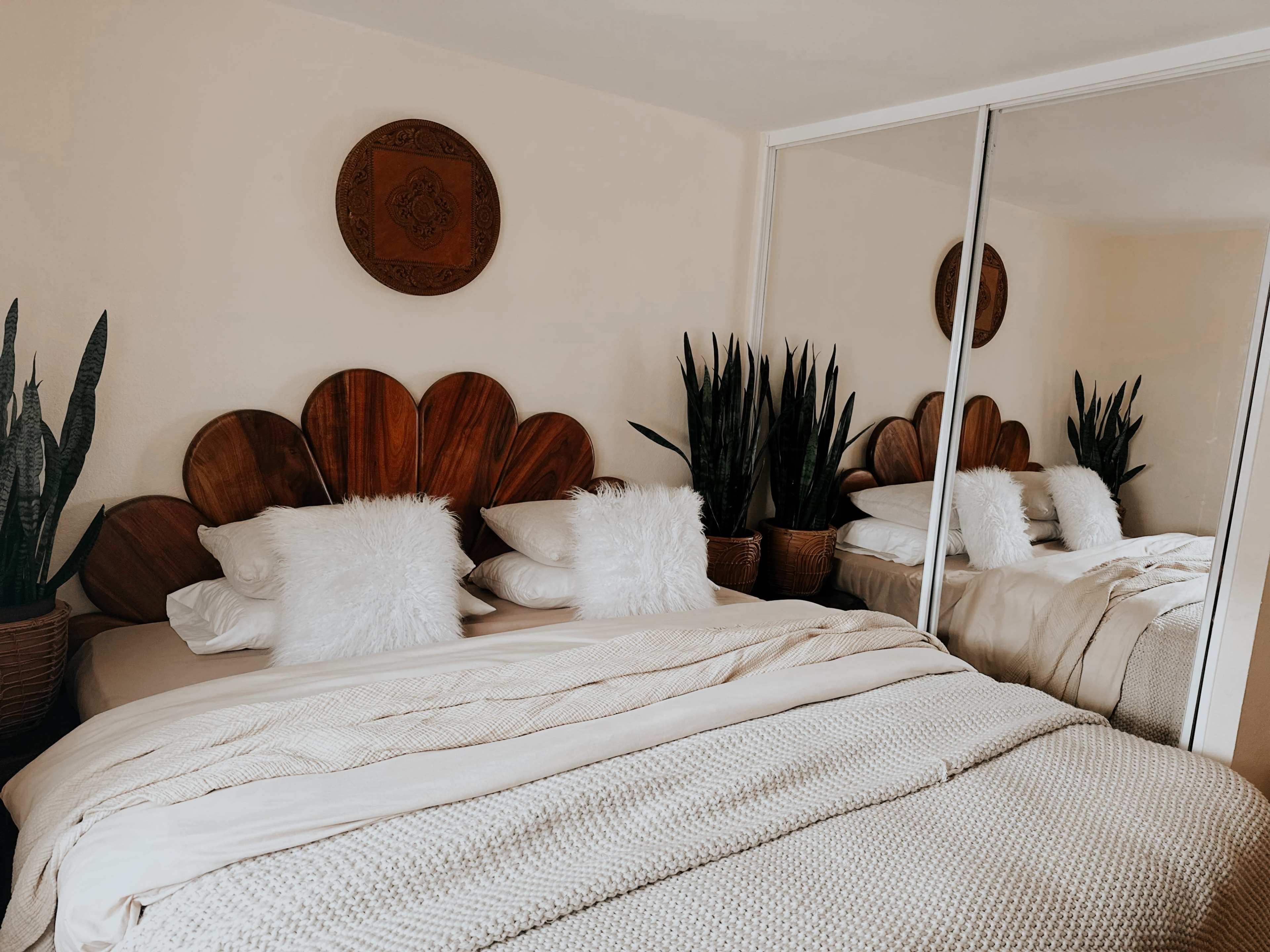 A neatly arranged bedroom with a bed featuring a decorative wooden headboard, two fluffy pillows, and plants in pots positioned beside mirrors on the wall.