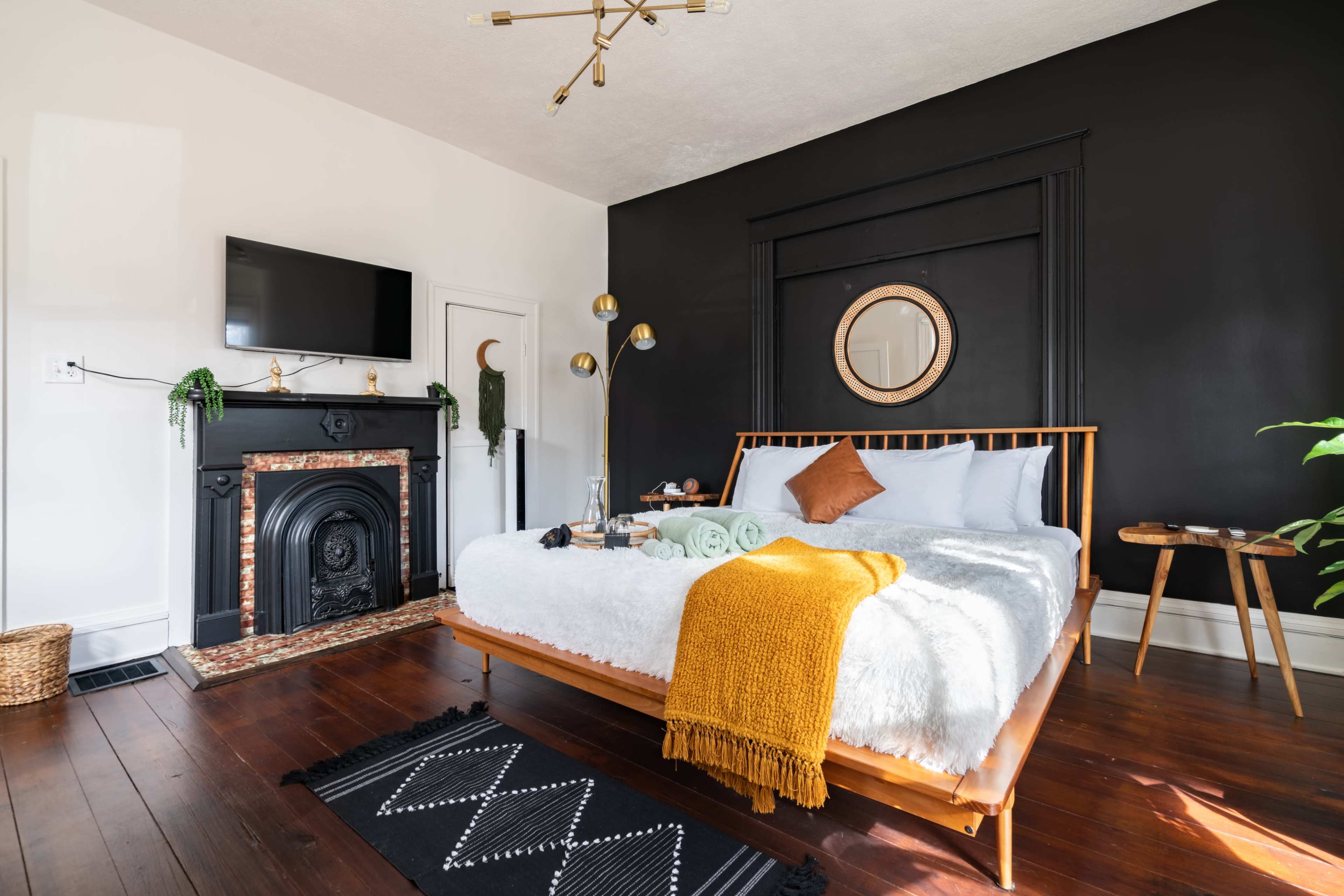 A modern bedroom featuring a wooden bed with white bedding, a black accent wall, a flat-screen TV, and a decorative fireplace.